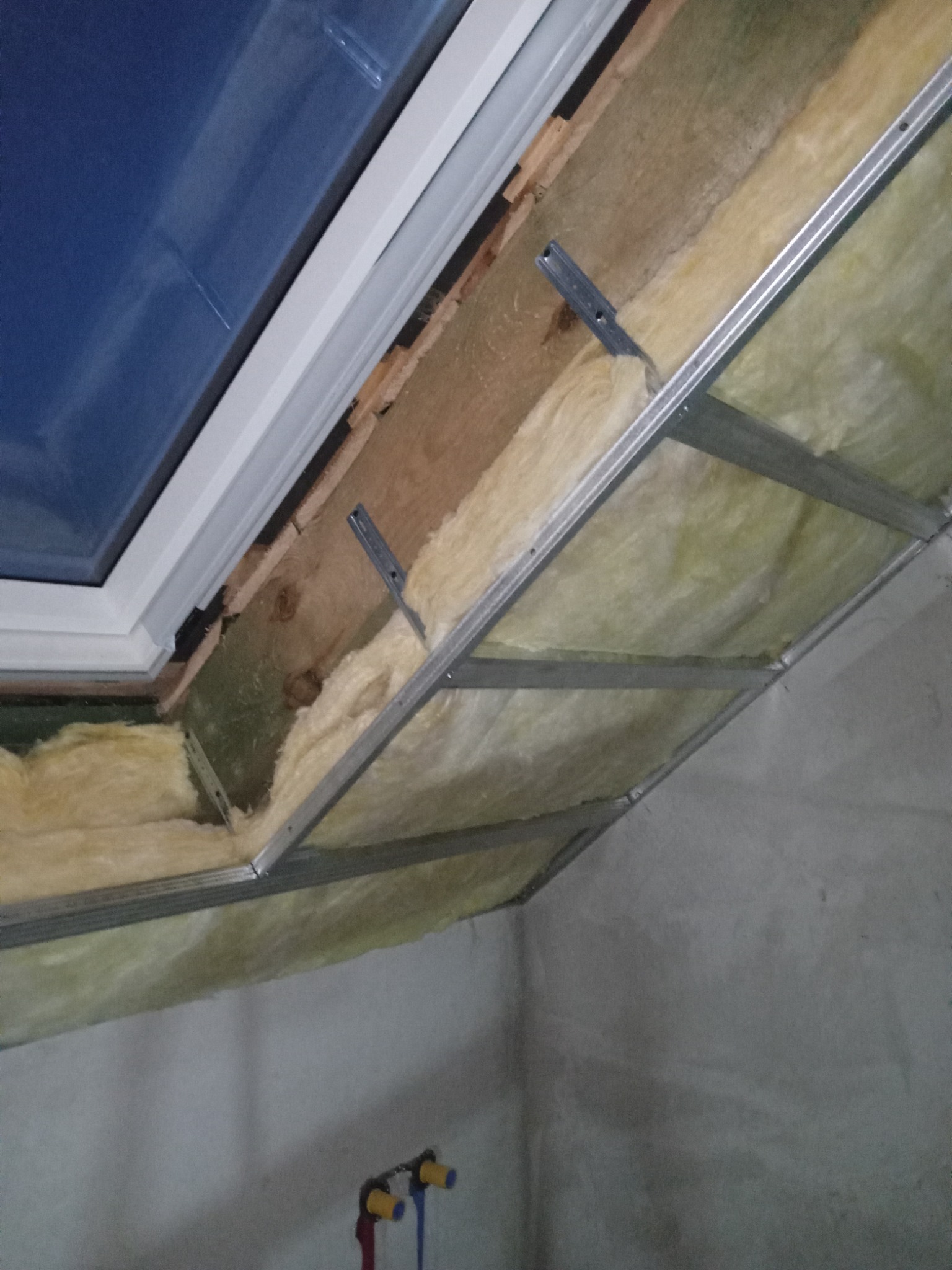 Montaż izolacji z wełny mineralnej i metalowej konstrukcji pod płytę karton gips wokół okna dachowego, z widocznymi elementami instalacji wodnej w tle.