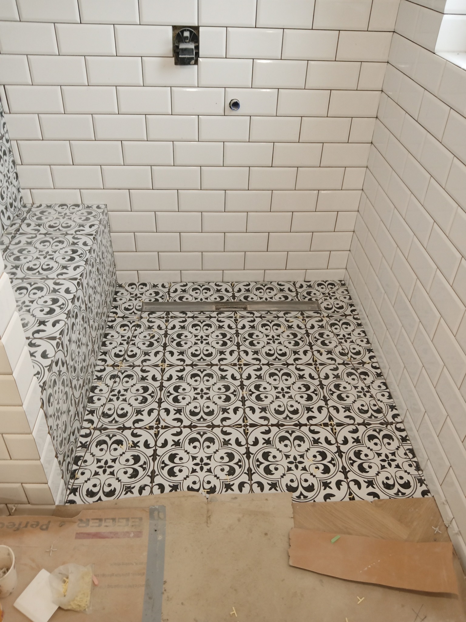 Wykończona kabina prysznicowa z białą, prostokątną glazurą ścienną i podłogą z czarno-białą, wzorzystą terakotą; widoczny odpływ liniowy i fragment zabudowanego siedziska.