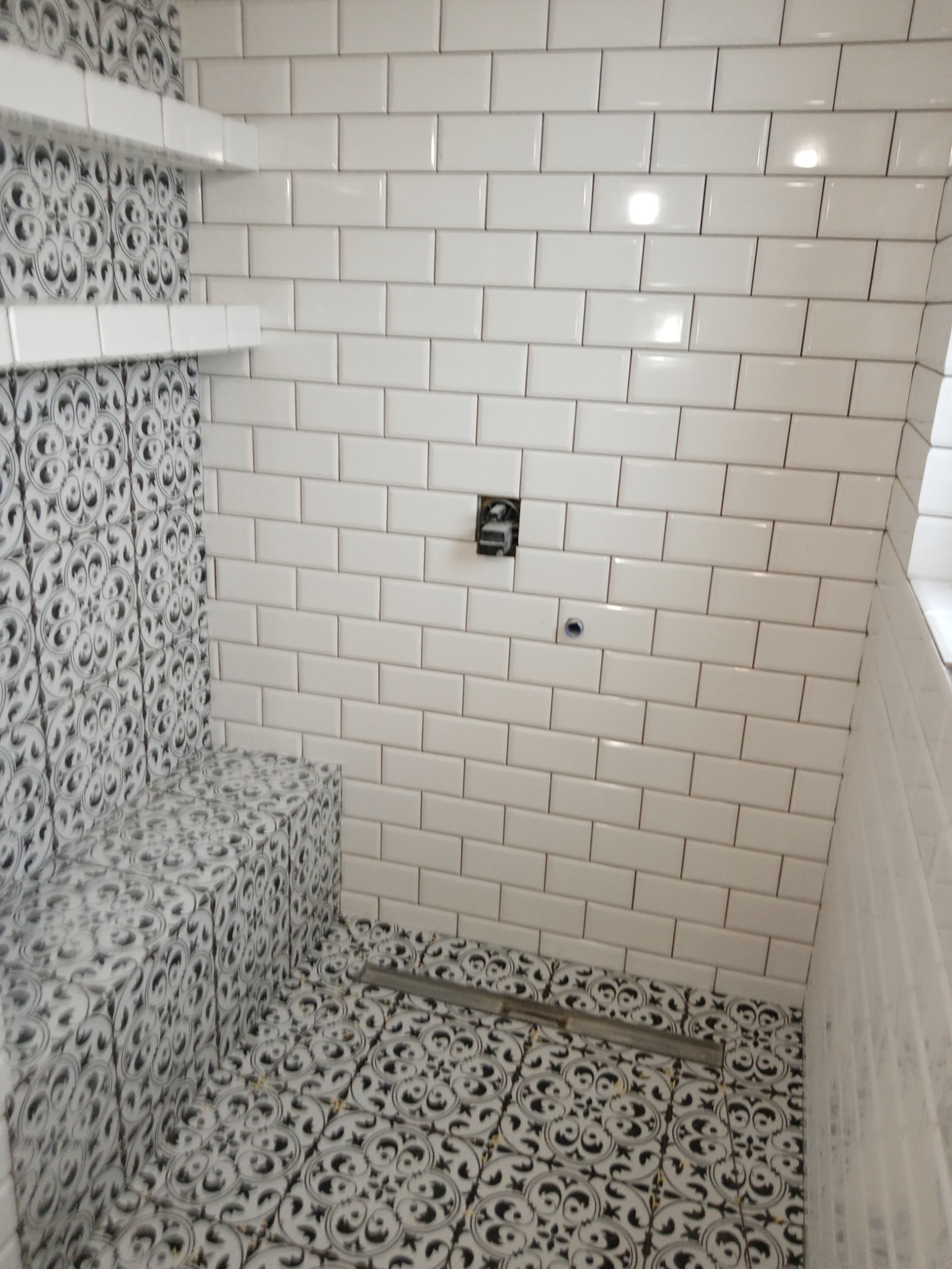 Wykończona kabina prysznicowa z białymi prostokątnymi płytkami na ścianach i dekoracyjnymi płytkami w kolorze czarno-białym na podłodze i siedzisku, widoczny odpływ liniowy i wystająca instalacja...