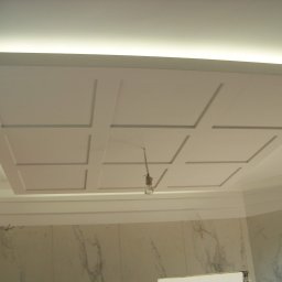 Dariusz Sadło - Kasetonowy sufit z podświetleniem LED i tymczasową żarówką w łazience z marmurowymi płytkami ściennymi.