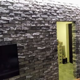 Dariusz Sadło - Ściana wykończona dekoracyjną cegłą w odcieniach szarości, z widocznym fragmentem telewizora i przejściem do innego pomieszczenia z kartonami.