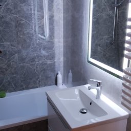 Dariusz Sadło - Nowoczesna łazienka z szarymi marmurkowymi płytkami, podświetlanym lustrem, białą umywalką z szafką i wanną z zielonym akcentem, minimalistyczny design.