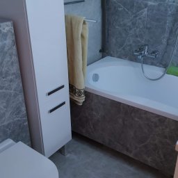 Dariusz Sadło - Wykończona łazienka z szarymi marmurkowymi płytkami, białą wanną, toaletą podwieszaną i szafką łazienkową, widok na baterię wannową i ręcznik na grzejniku.