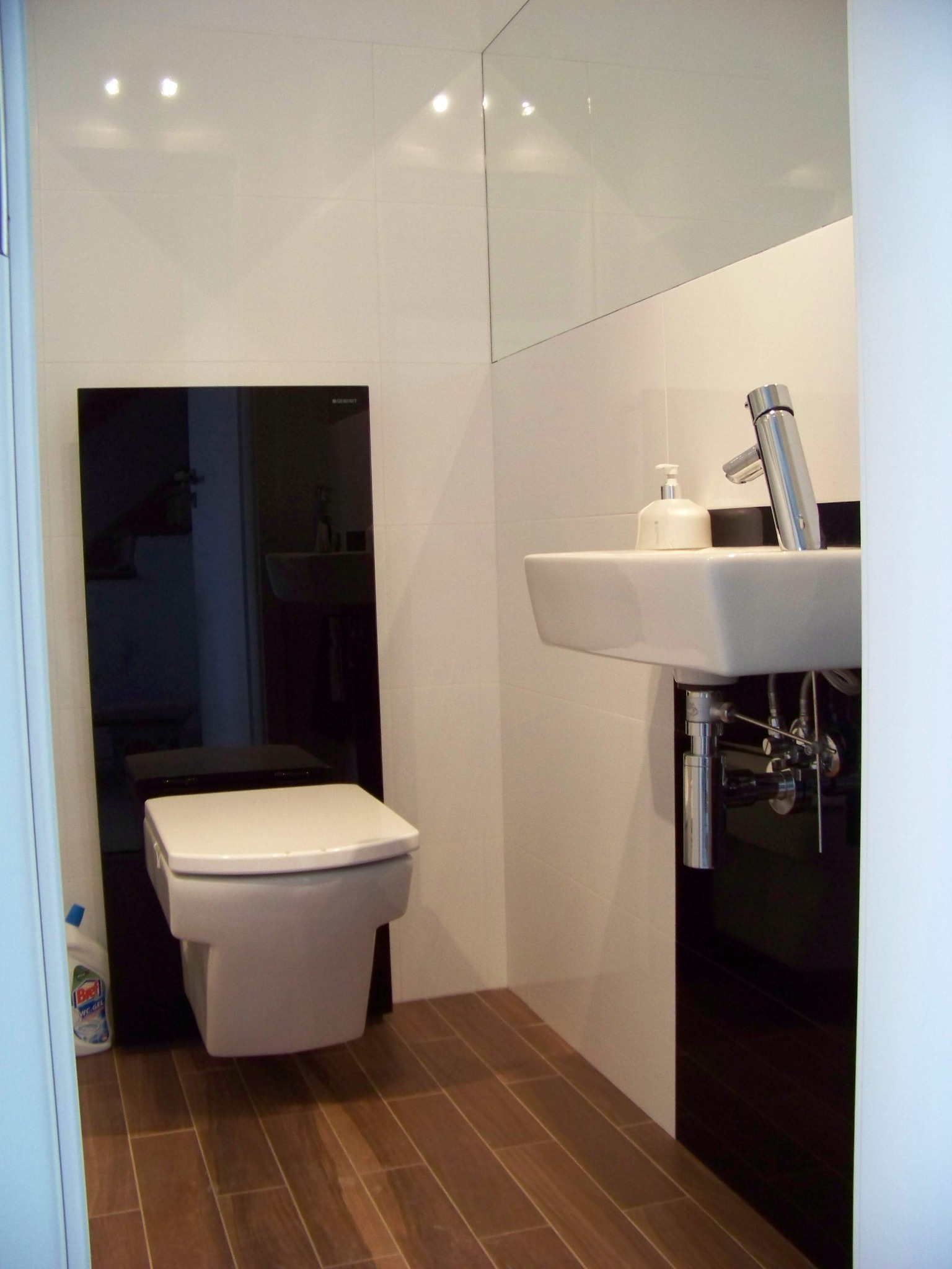 Nowoczesna łazienka w Częstochowie z podwieszaną toaletą Geberit na czarnym tle, białą umywalką z chromowaną baterią i drewnopodobną podłogą, oświetlona punktowymi lampami.