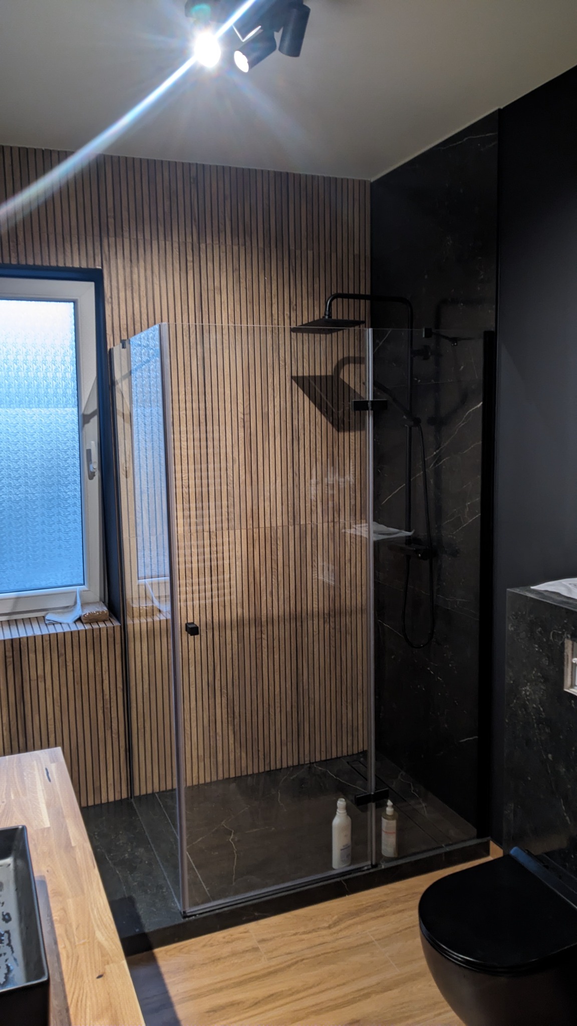 Nowoczesna łazienka z kabiną prysznicową typu walk-in, drewnianymi panelami ściennymi i czarnymi elementami armatury, widoczne butelki z kosmetykami wewnątrz kabiny.