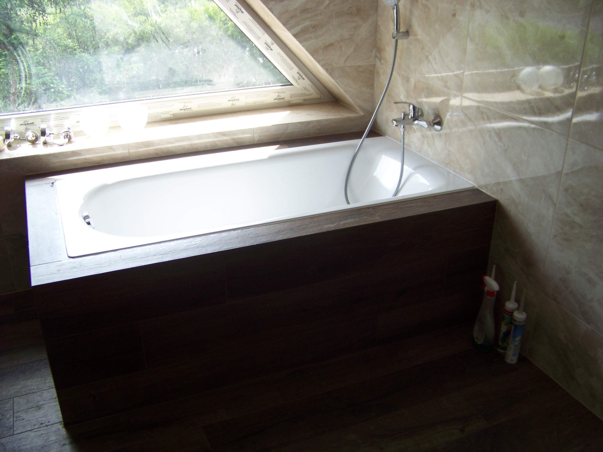 Jasna łazienka z wanną obudowaną ciemnym drewnem, okno dachowe z widokiem na zieleń, marmurowe płytki na ścianach, ciemna podłoga, widoczne akcesoria łazienkowe i środki czystości w rogu.