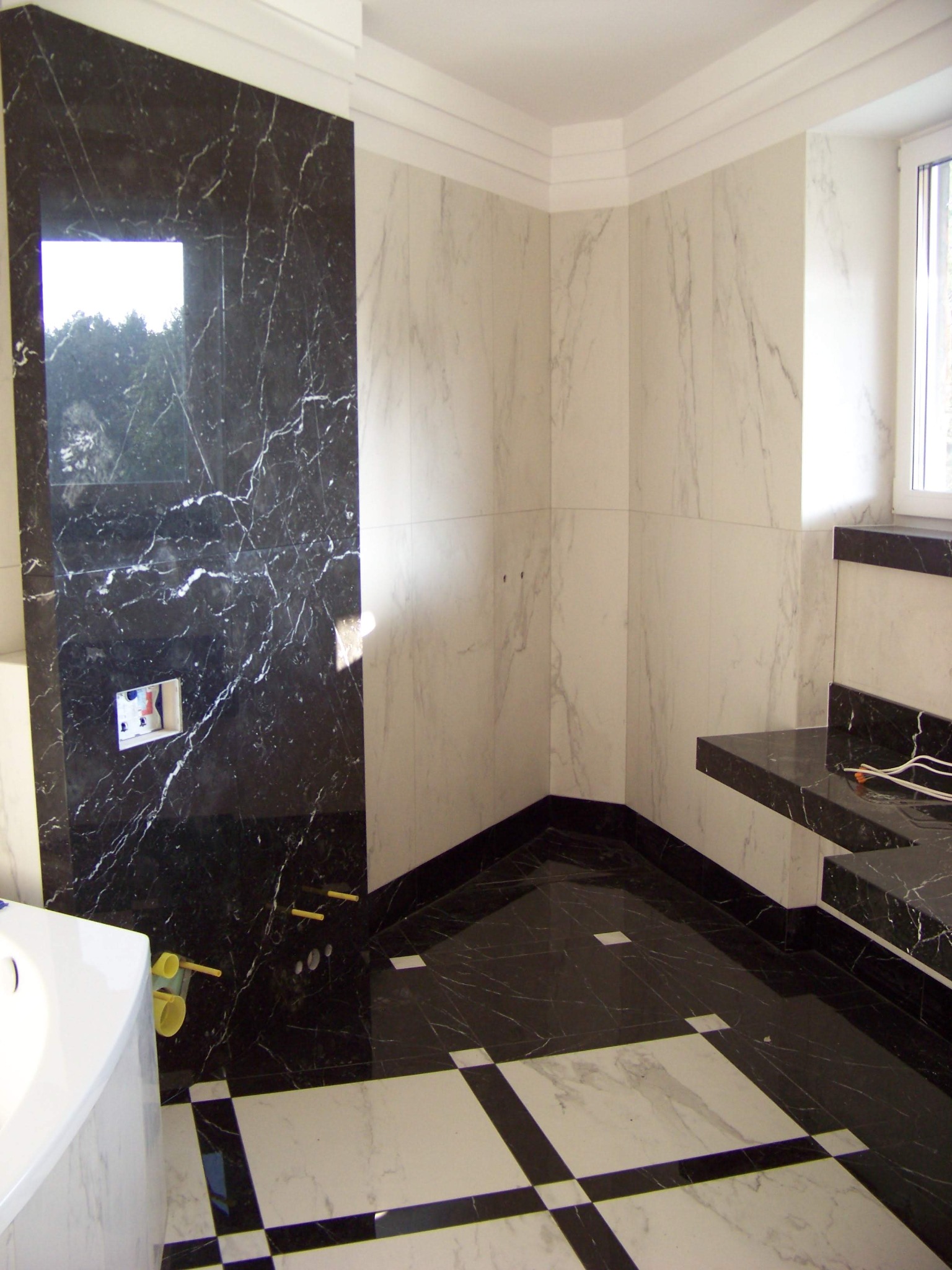 Wnętrze łazienki w trakcie remontu: ściany wyłożone białymi i czarnymi marmurowymi płytami, podłoga w szachownicę, widoczne instalacje hydrauliczne i elektryczne, brak armatury.