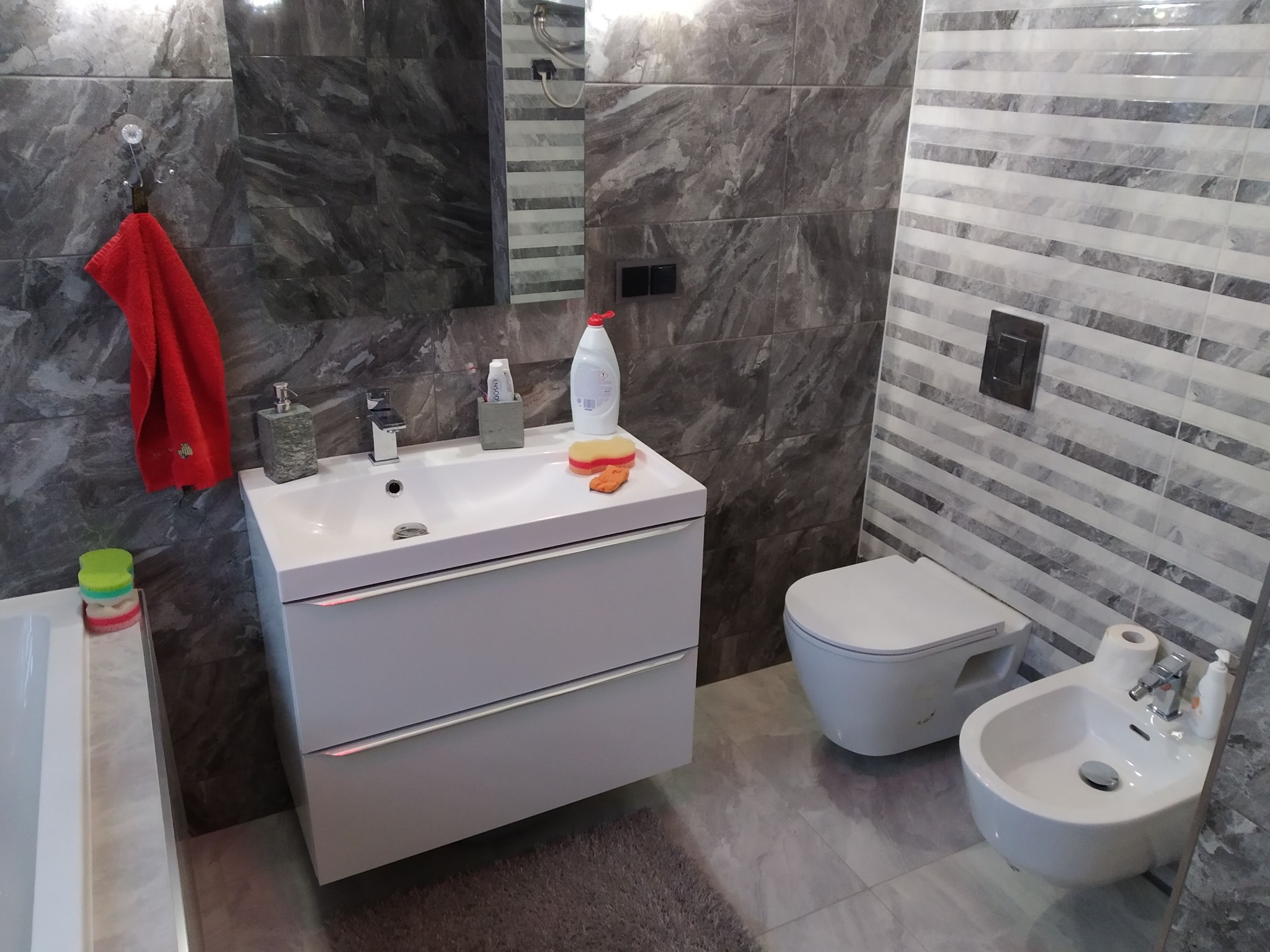 Nowoczesna łazienka z szarymi płytkami imitującymi marmur, białą szafką pod umywalką z szufladami, wiszącą toaletą i bidetem. Na umywalce leży gąbka i płyn do naczyń, a na ścianie wisi czerwony...