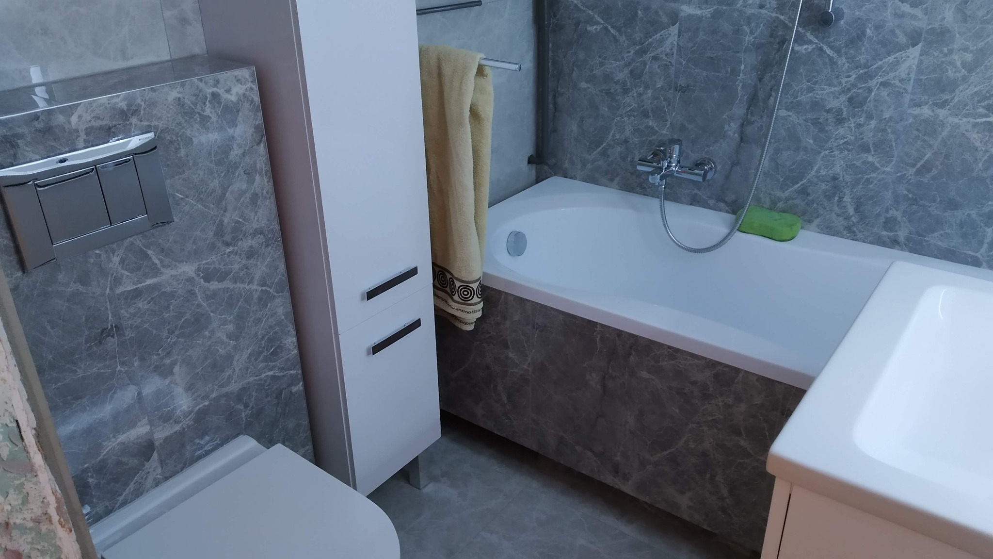 Wykończona łazienka z szarymi marmurkowymi płytkami, białą wanną, toaletą podwieszaną i szafką łazienkową, widok na baterię wannową i ręcznik na grzejniku.