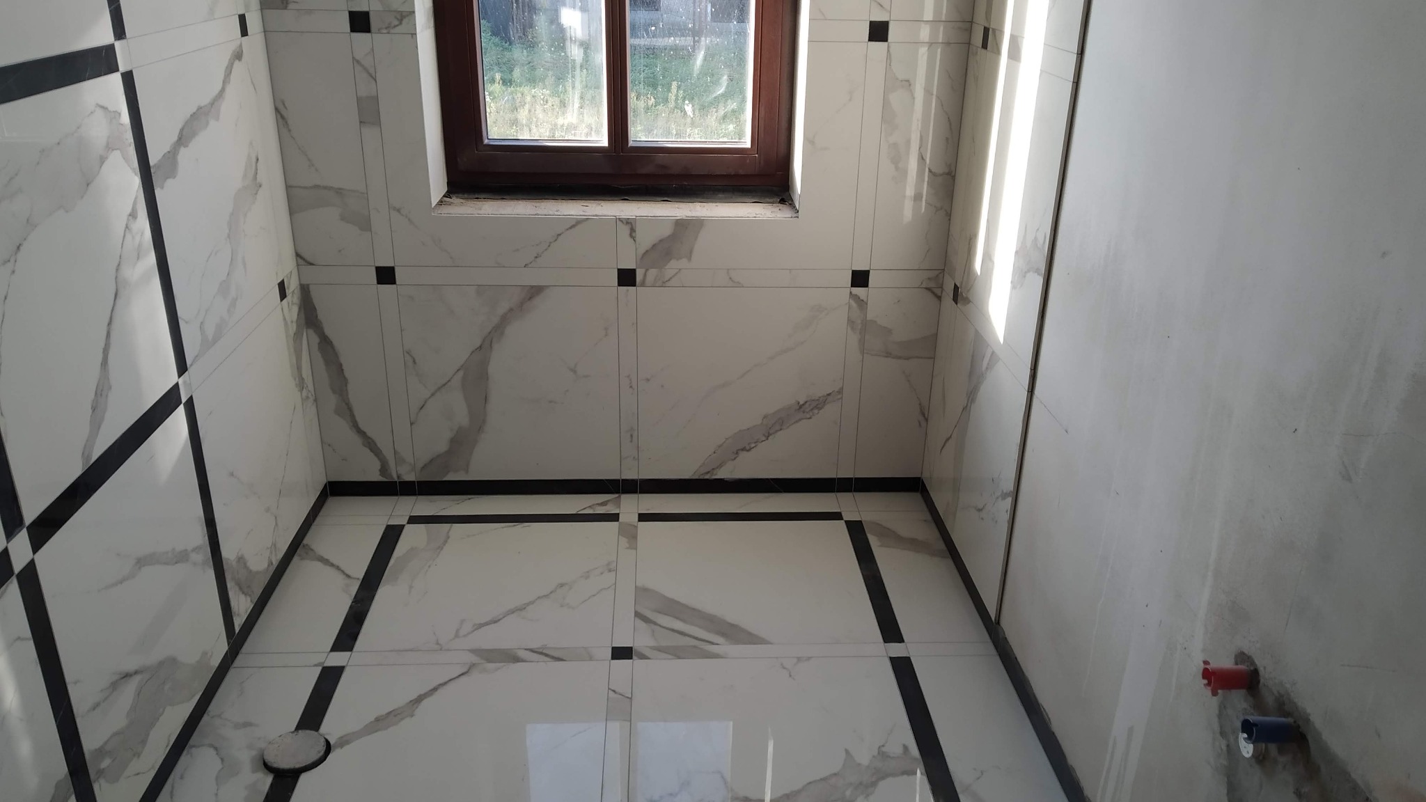 Wykończona łazienka z białymi płytkami imitującymi marmur, czarne akcenty na fugach i listwach, drewniane okno w tle, widoczne przyłącza wodne na surowej ścianie.