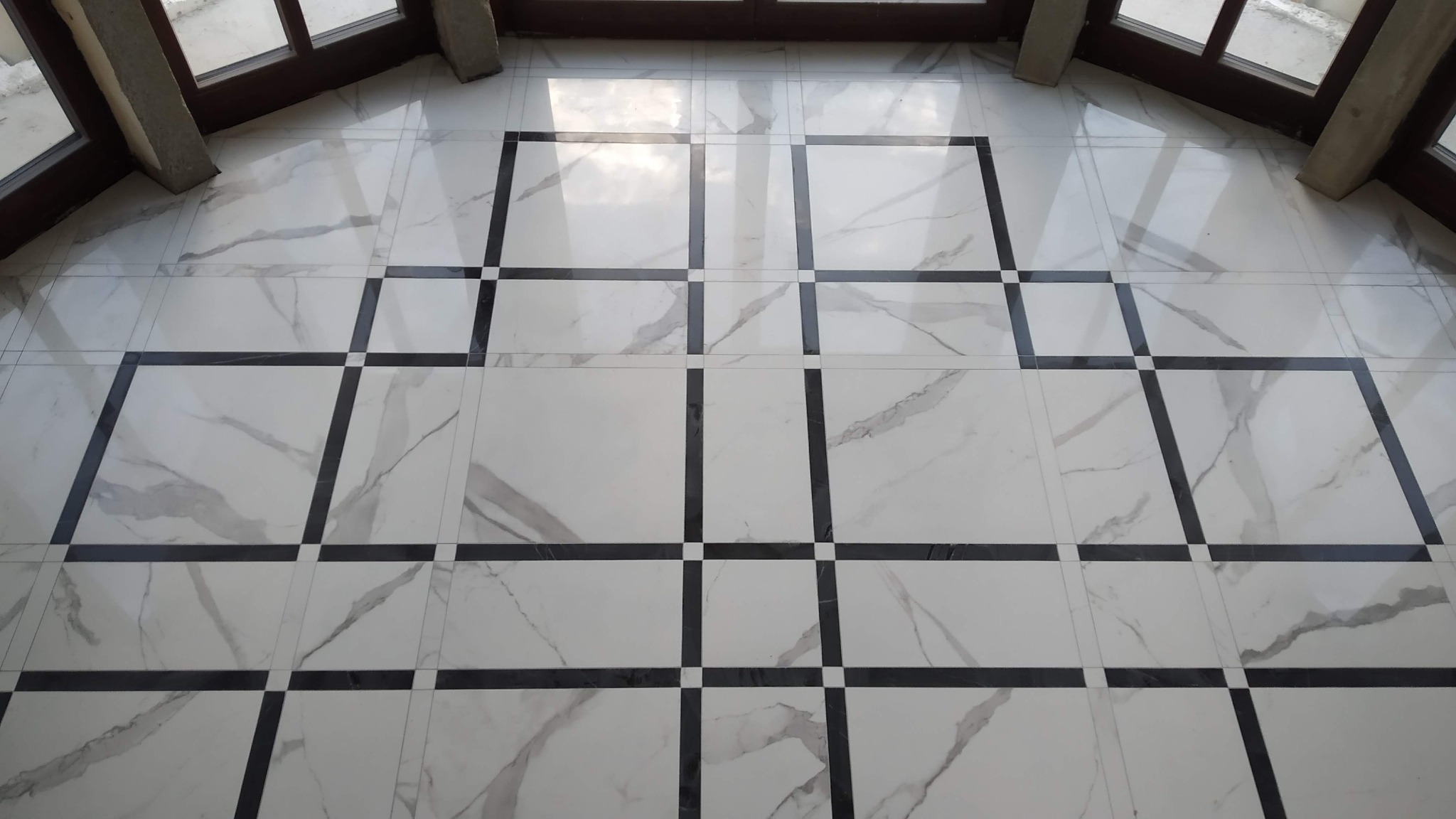Białe marmurowe płytki podłogowe z czarnymi wstawkami tworzącymi geometryczny wzór kwadratów, widoczne z góry, z oknem w tle.