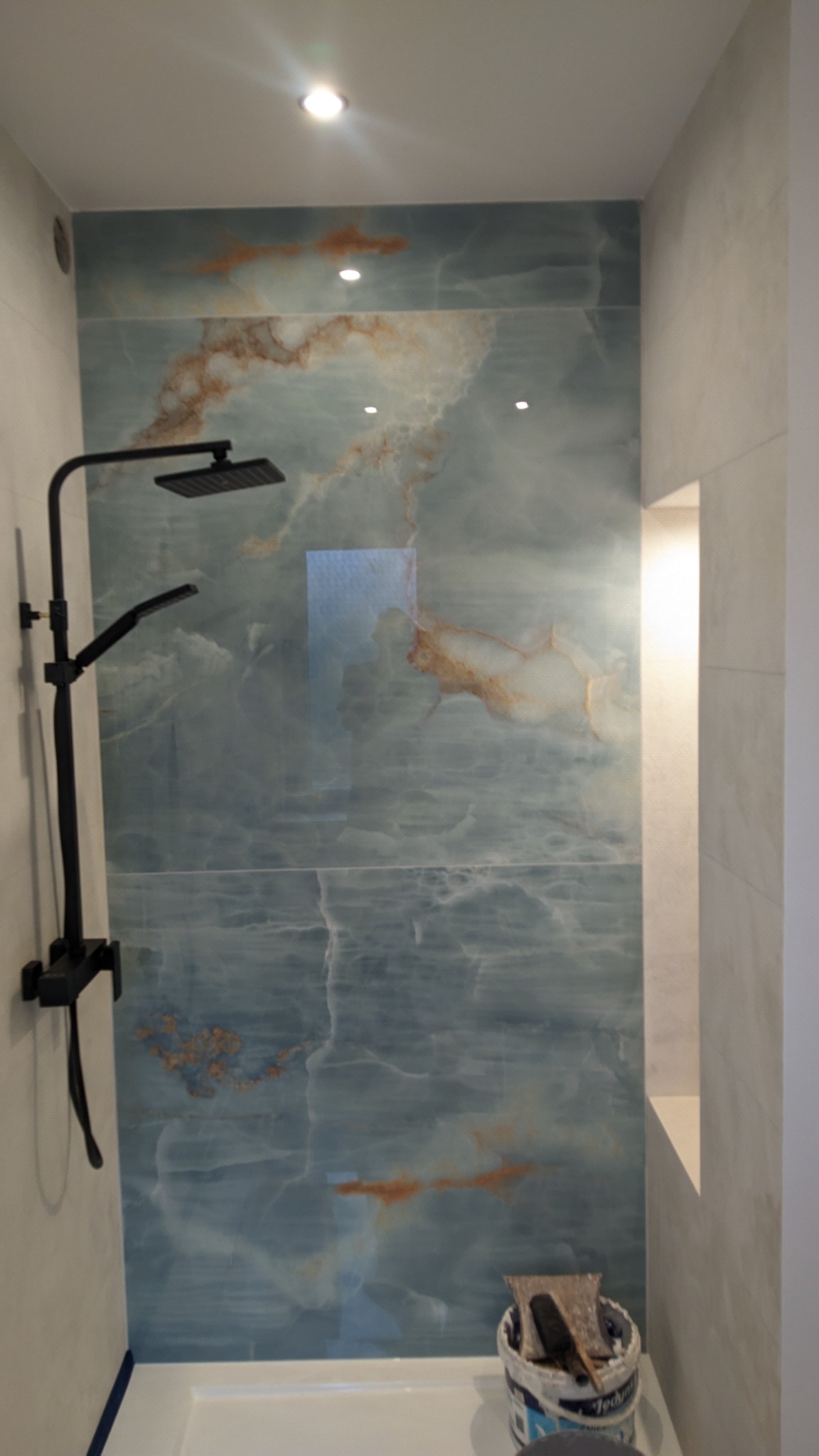 Nowoczesna łazienka z kabiną prysznicową typu walk-in, wykończona płytami imitującymi onyks w odcieniach błękitu i brązu, czarny zestaw natryskowy, wiadro z narzędziami na dnie kabiny.