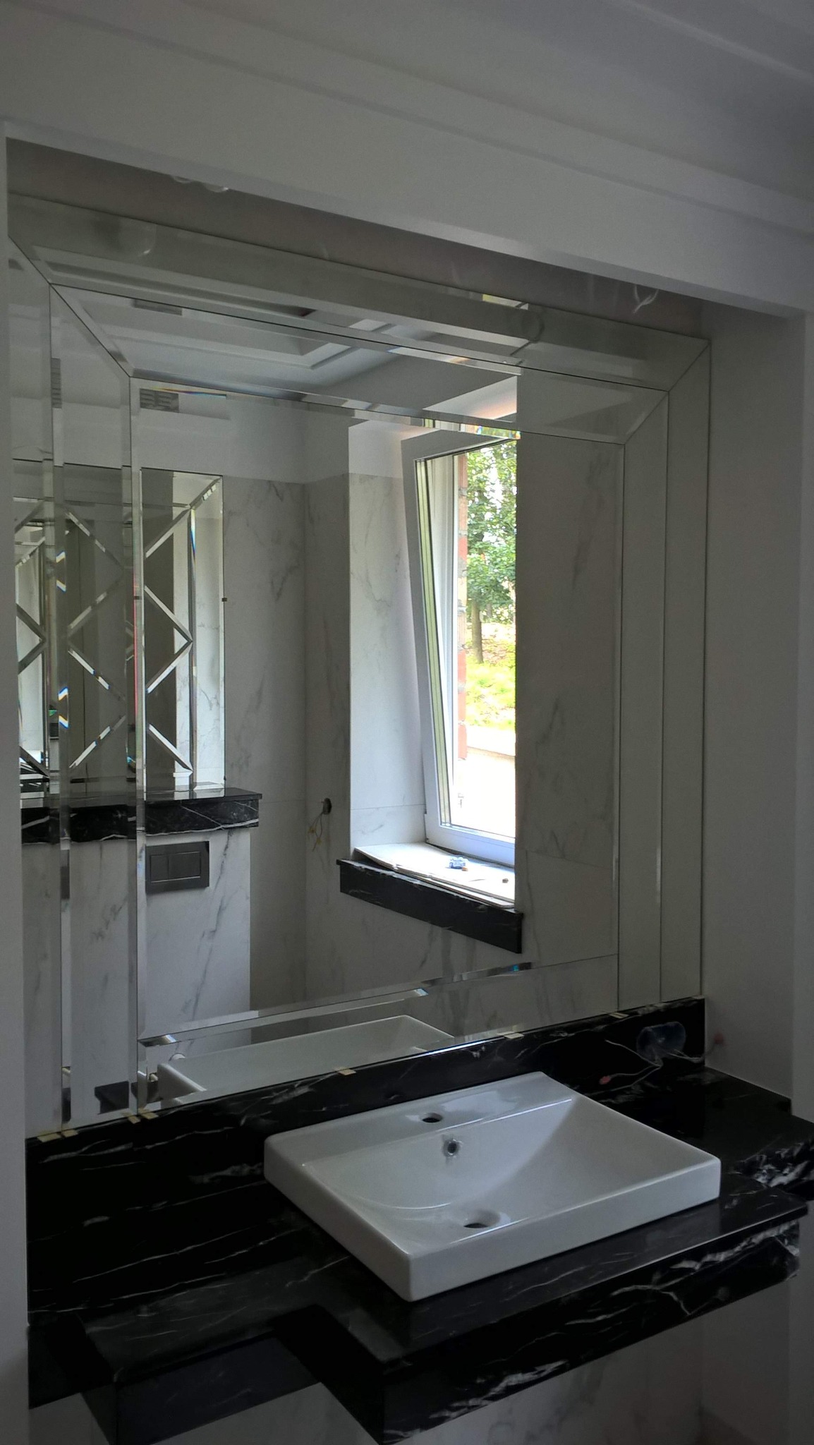 Nowoczesna łazienka z dużym lustrem z fazowanymi krawędziami, białą umywalką nablatową na czarnym blacie z marmuru oraz marmurowymi płytkami na ścianach.