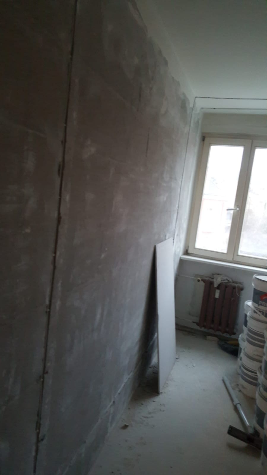 Ściana w trakcie szpachlowania z widocznymi łączeniami płyt gipsowo-kartonowych, fragment okna i grzejnika w tle, oraz stos farb w wiadrach po prawej stronie.