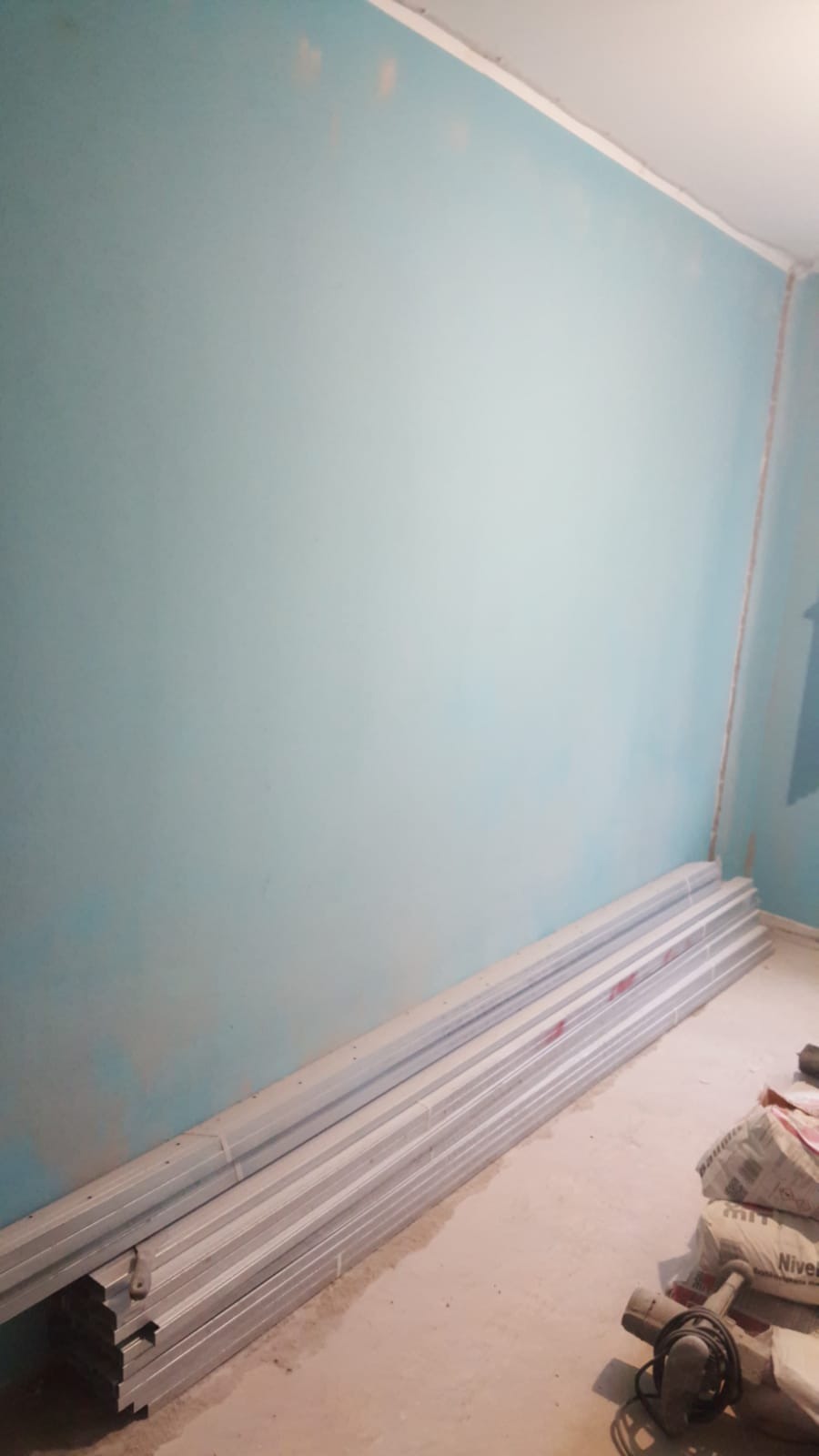 Niebieska ściana w trakcie przygotowania do prac wykończeniowych, widoczne profile aluminiowe, worki z zaprawą i szlifierka kątowa na podłodze.