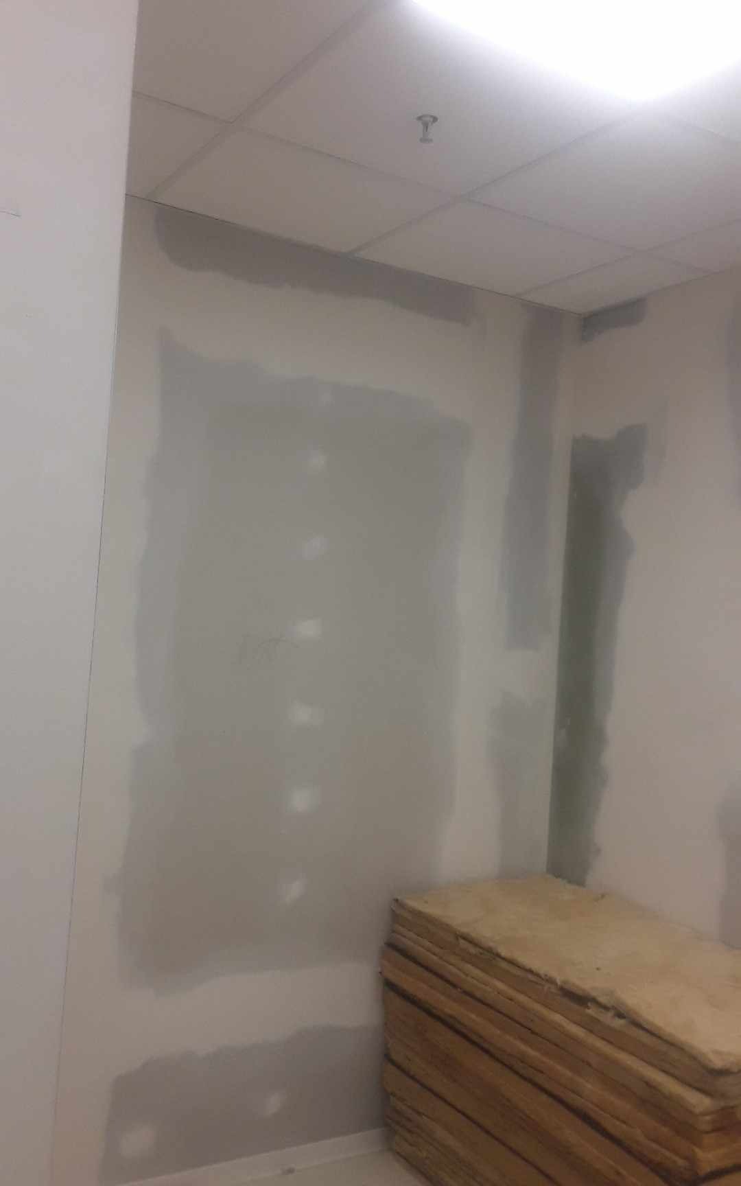 Ściana w trakcie szpachlowania z widocznymi punktami po wkrętach, obok stos płyt drewnianych, sufit podwieszany w tle.