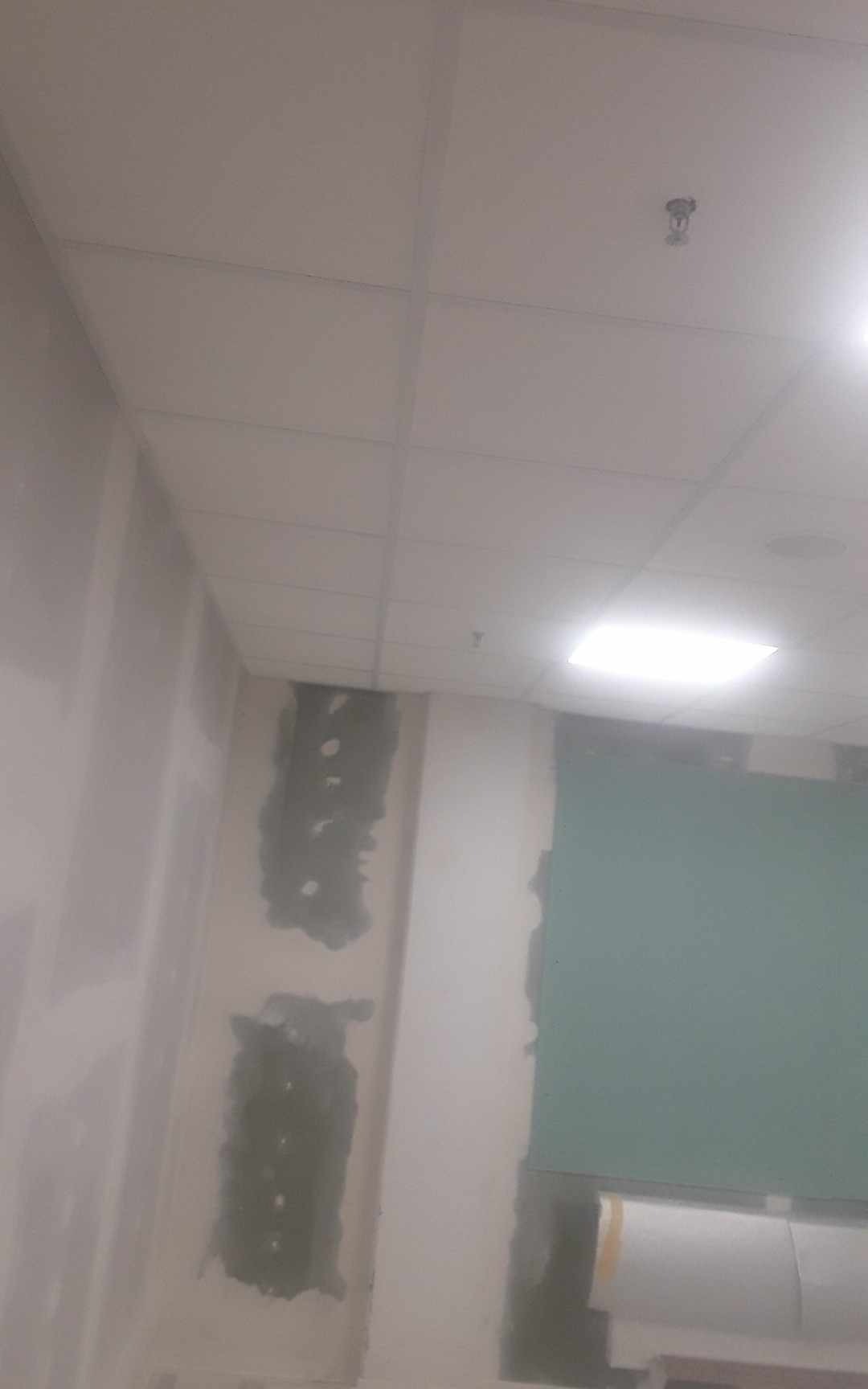 Wnętrze w trakcie prac wykończeniowych: sufit kasetonowy z widocznym systemem oświetlenia i czujnikiem dymu, ściany częściowo wyszpachlowane z widocznymi śladami po łatach, fragment zielonej ściany...