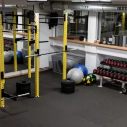 Wnętrze siłowni: żółte konstrukcje do ćwiczeń, sztangi, hantle na stojakach, piłki gimnastyczne, podłoga gumowa.