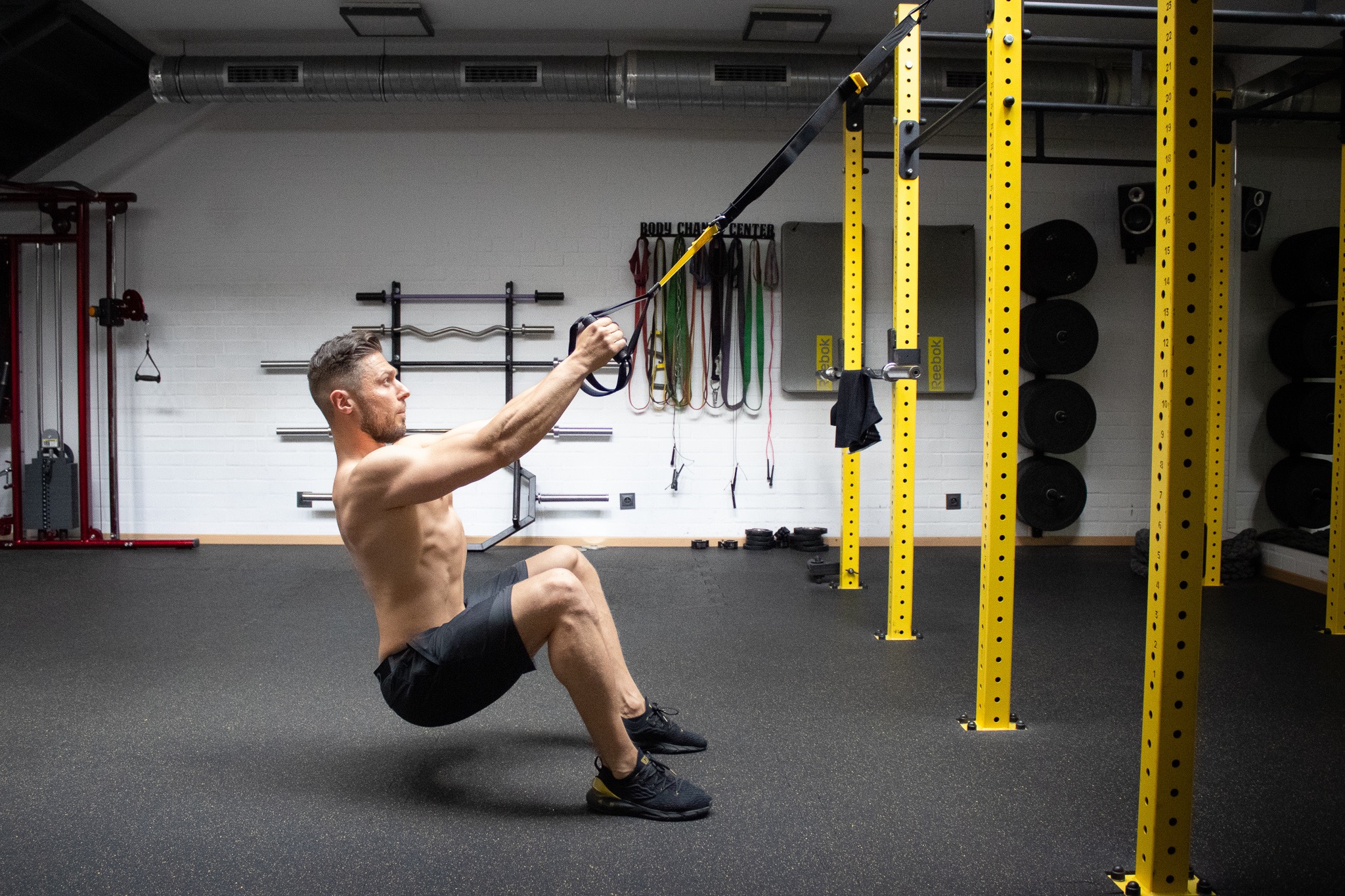 Mężczyzna w szortach wykonuje ćwiczenie na TRX w siłowni, skupiony wzrok, widoczny sprzęt do ćwiczeń siłowych i akcesoria zawieszone na ścianie.