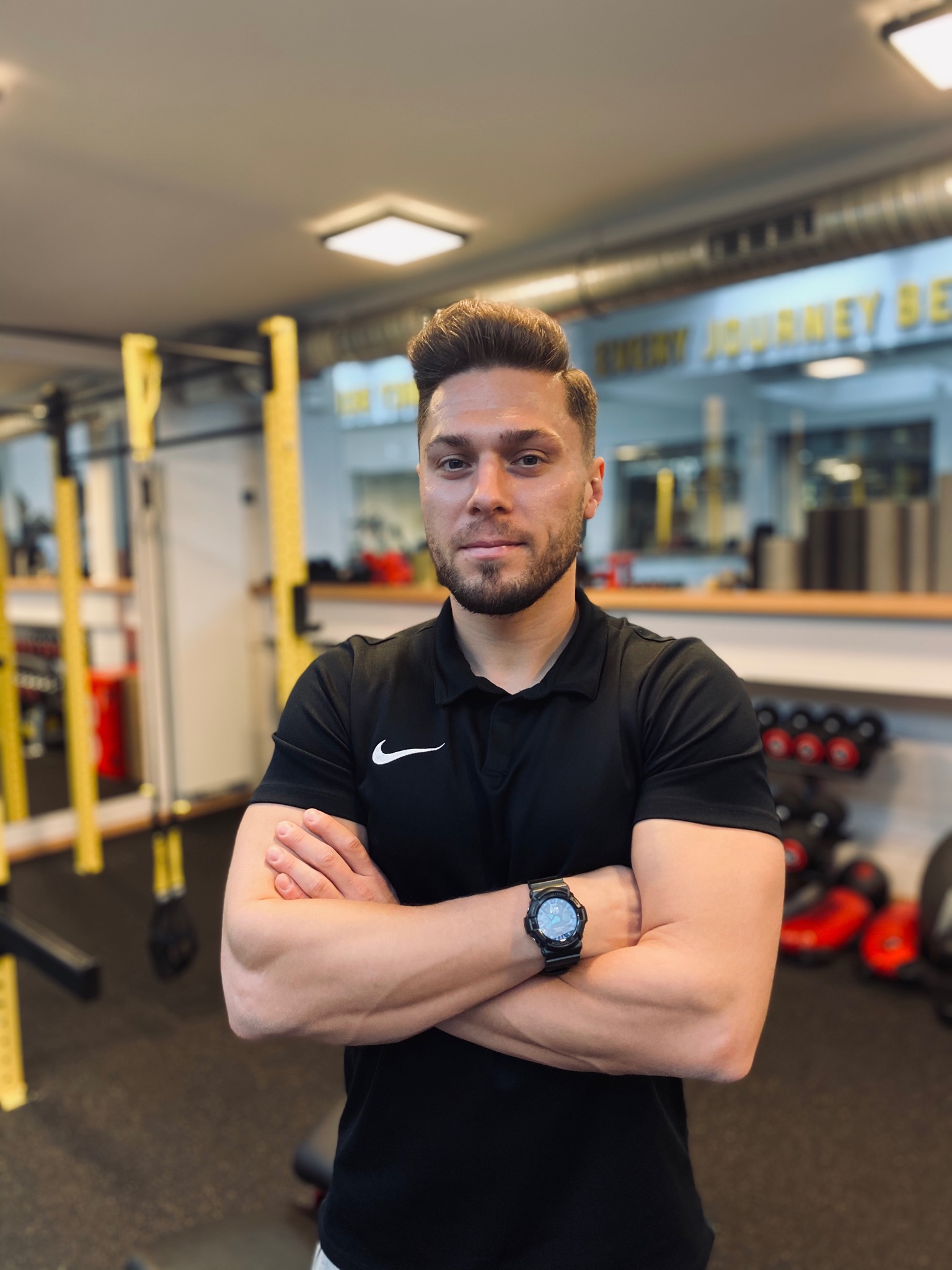 Portret mężczyzny w czarnej koszulce polo z logo Nike, stojącego z założonymi rękami na tle siłowni z żółtymi elementami konstrukcyjnymi i sprzętem do ćwiczeń.