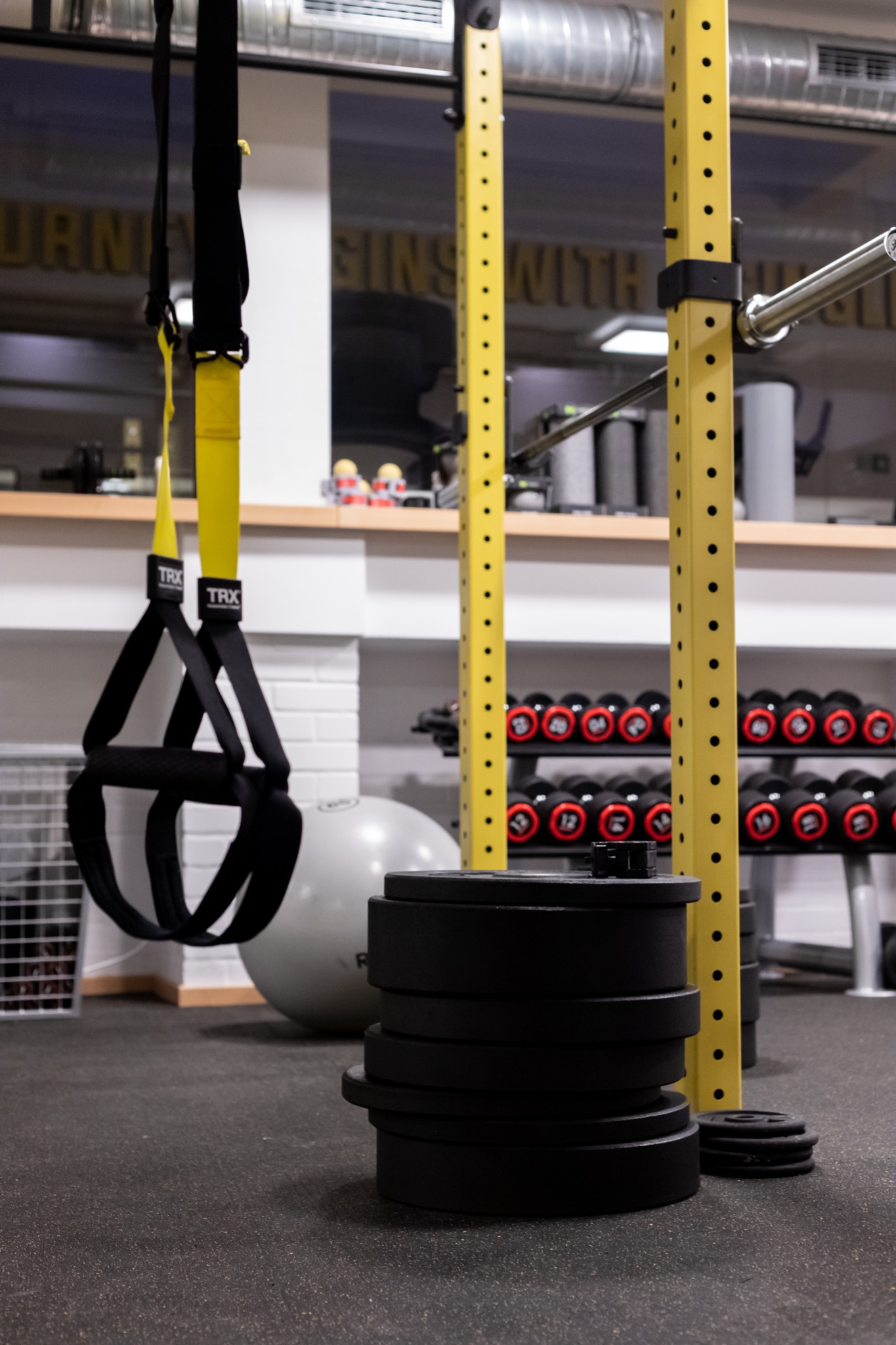 Wyposażenie siłowni: czarne obciążenia na podłodze, żółta klatka do ćwiczeń, zestaw hantli i zawieszone pasy TRX.