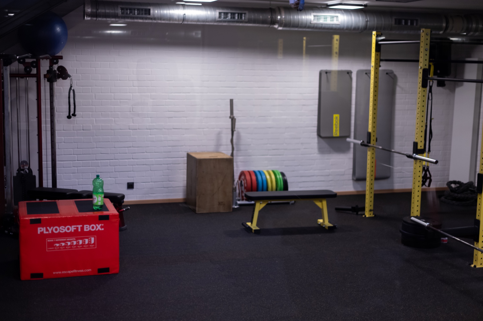 Wnętrze siłowni z wyposażeniem: skrzynia plyometryczna z butelką wody, ławka do ćwiczeń, stojak na obciążenia, drabinki, gumy do ćwiczeń i piłka gimnastyczna.