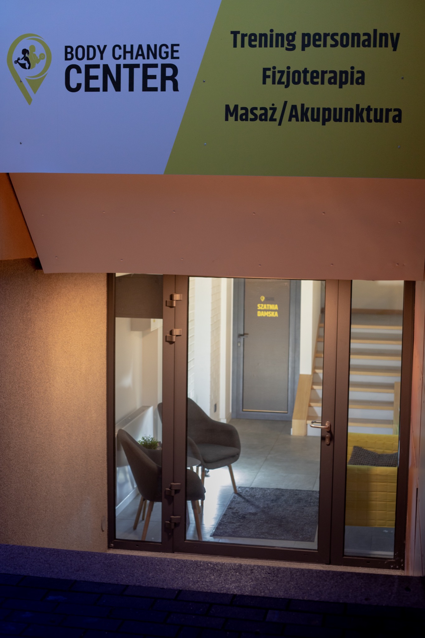 Wejście do Body Change Center z widocznym logo, ofertą usług (trening personalny, fizjoterapia, masaż/akupunktura) i wnętrzem z krzesłami oraz schodami.