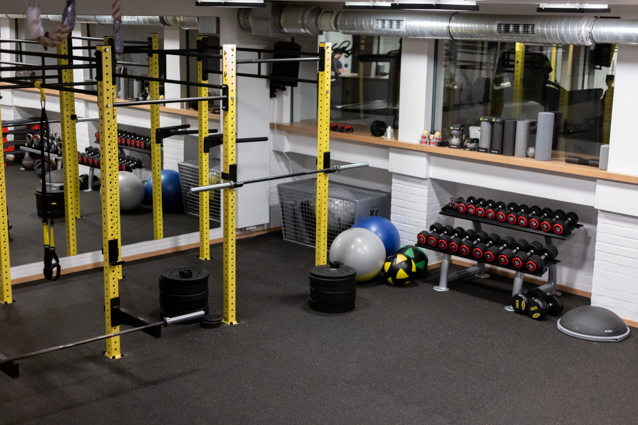 Wnętrze siłowni: żółte konstrukcje do ćwiczeń, sztangi, hantle na stojakach, piłki gimnastyczne, podłoga gumowa.