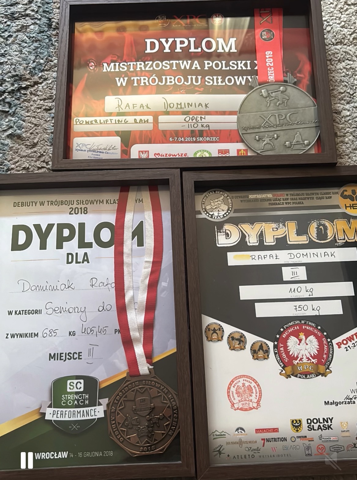 Dyplomy i medale z zawodów trójboju siłowego Rafała Dominiaka, w tym medal z Mistrzostw Polski XPC, debiutów w trójboju siłowym oraz inne.