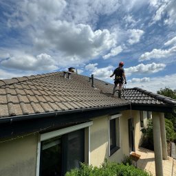 Loba FrogForce - Pracownik w szelkach asekuracyjnych czyści dach z brązowej dachówki ceramicznej, widoczne dwa kominy wentylacyjne i błękitne niebo z obłokami.