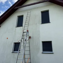 Mycie elewacji domu: widoczny efekt czyszczenia na ścianie, drabina oparta o budynek, zamknięte rolety okienne, błękitne niebo z chmurami w tle.