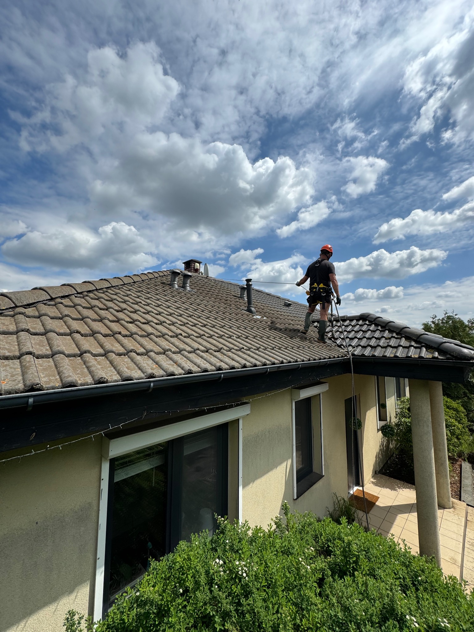 Pracownik w szelkach asekuracyjnych czyści dach z brązowej dachówki ceramicznej, widoczne dwa kominy wentylacyjne i błękitne niebo z obłokami.