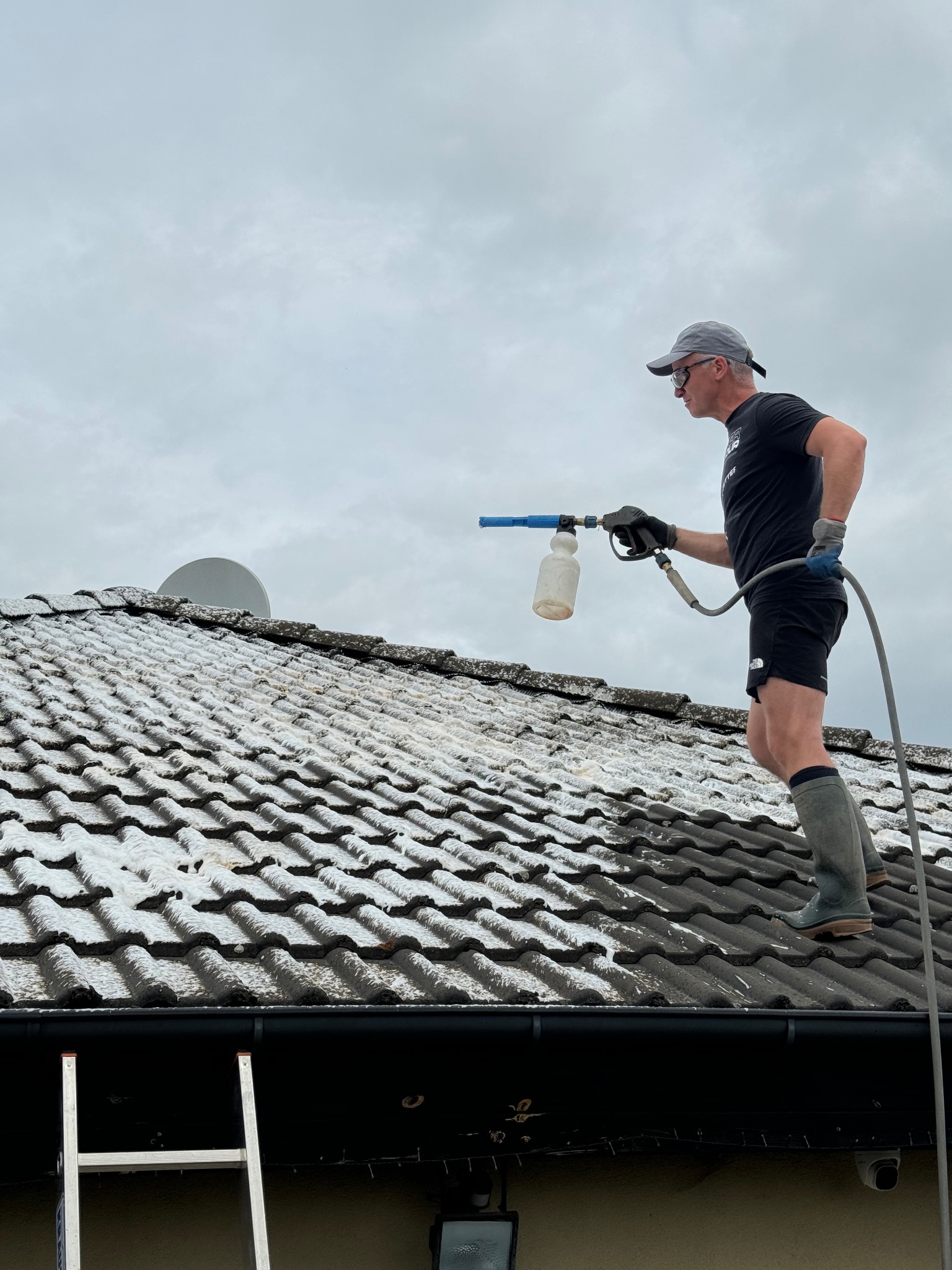 Mężczyzna w okularach i czapce, stojący na dachu pokrytym brudem i mchem, czyści go za pomocą myjki ciśnieniowej z pojemnikiem na detergent; w tle pochmurne niebo i antena satelitarna.