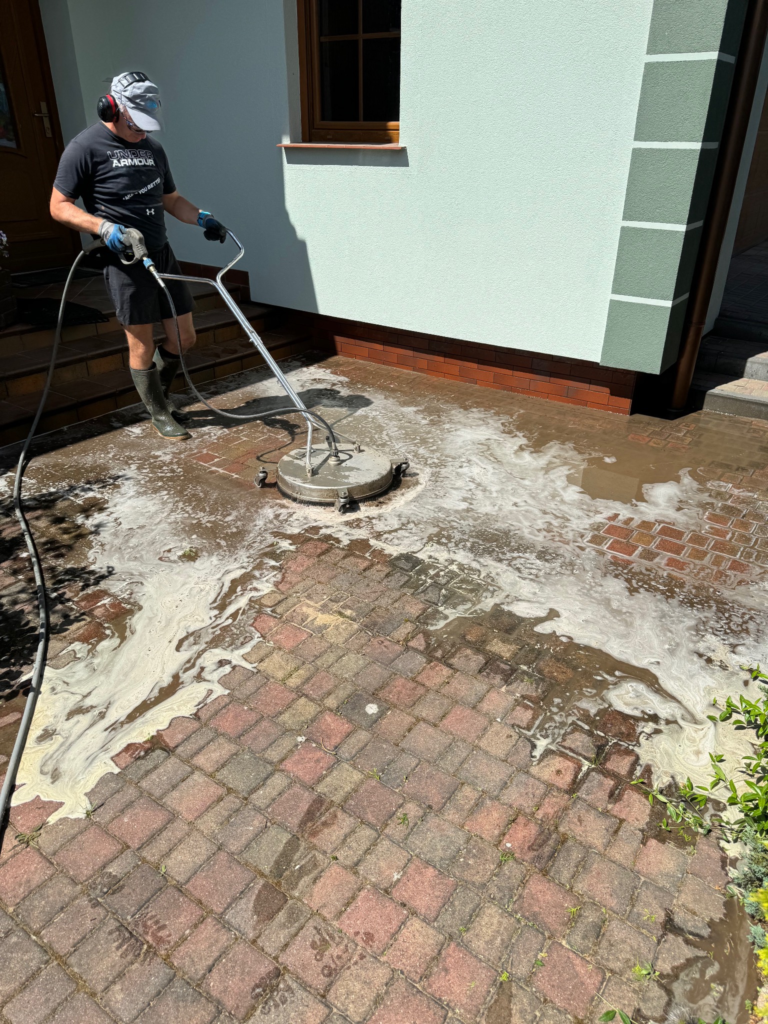 Mężczyzna w gumowych butach czyści czerwoną kostkę brukową za pomocą myjki ciśnieniowej z okrągłą dyszą, w tle jasna elewacja domu z brązowymi akcentami.