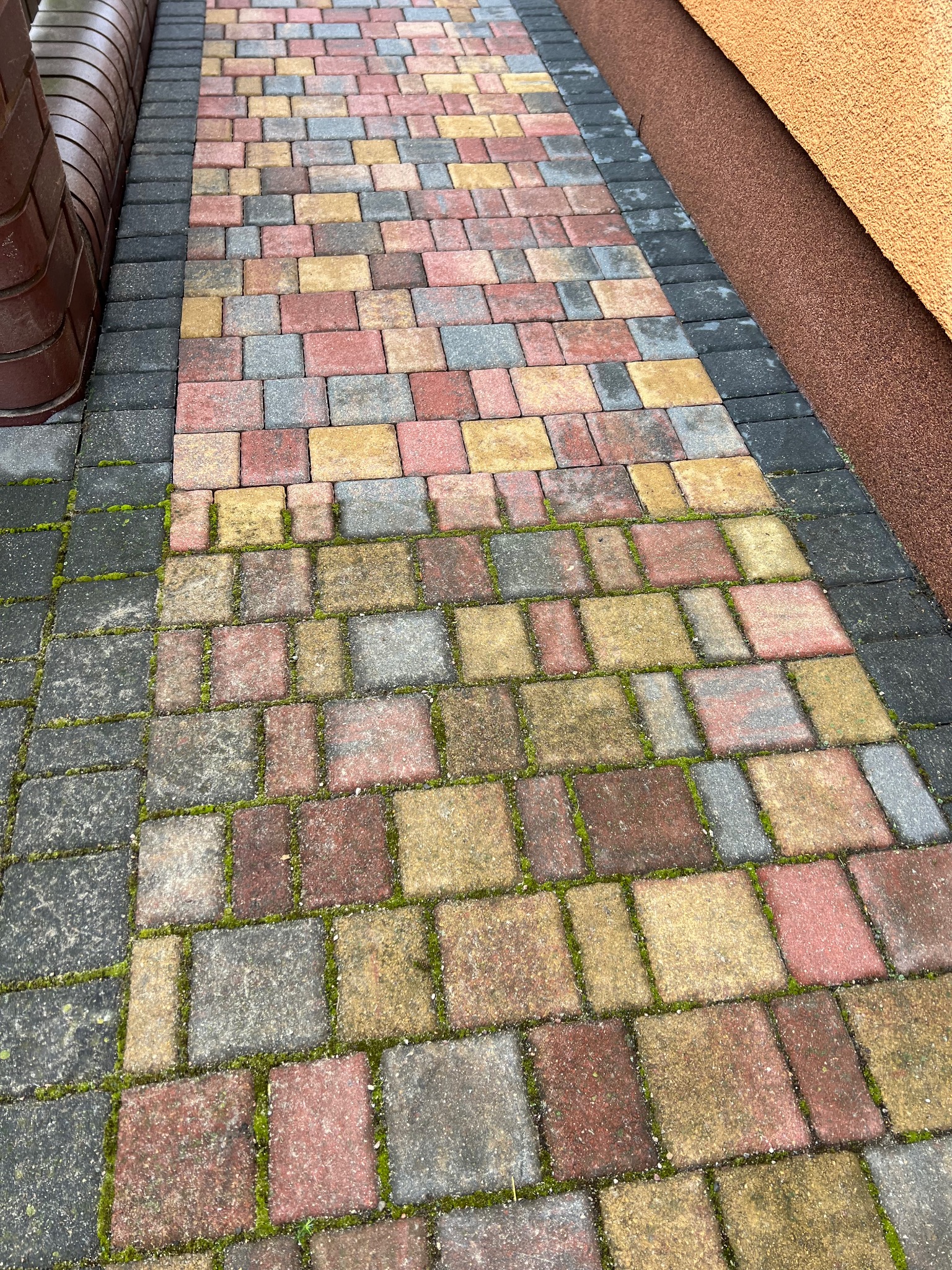 Kolorowa kostka brukowa ułożona wzdłuż ściany budynku, z widocznym mchem w spoinach.