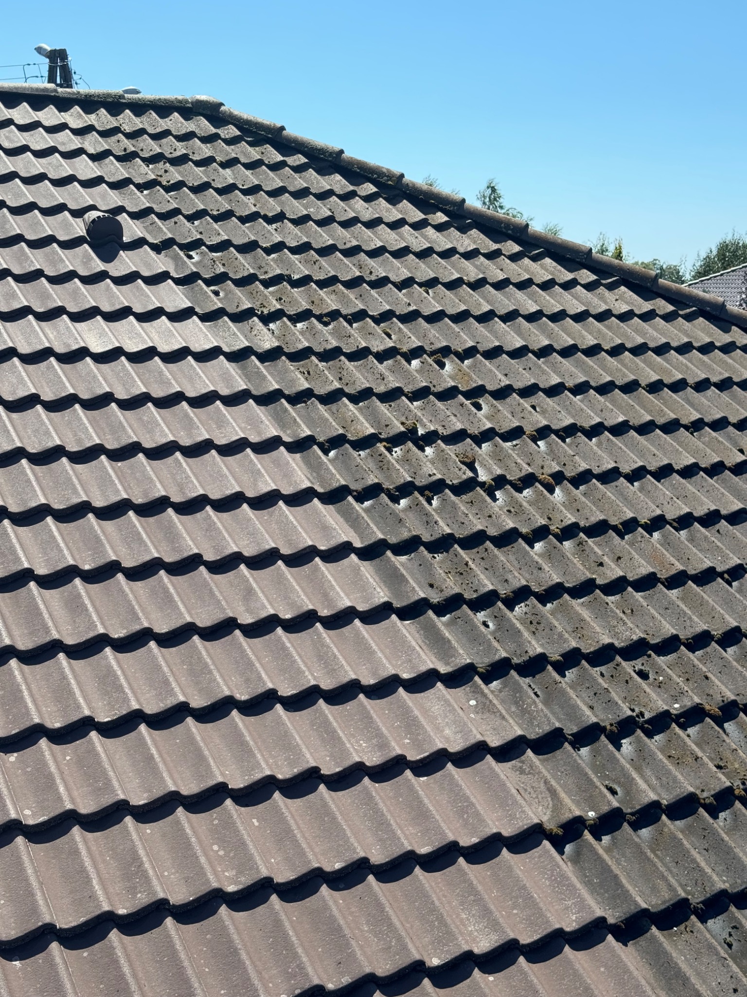 Ciemnobrązowa dachówka ceramiczna pokryta mchem i zabrudzeniami, widok z góry na fragment dachu z kominem wentylacyjnym, błękitne niebo w tle.