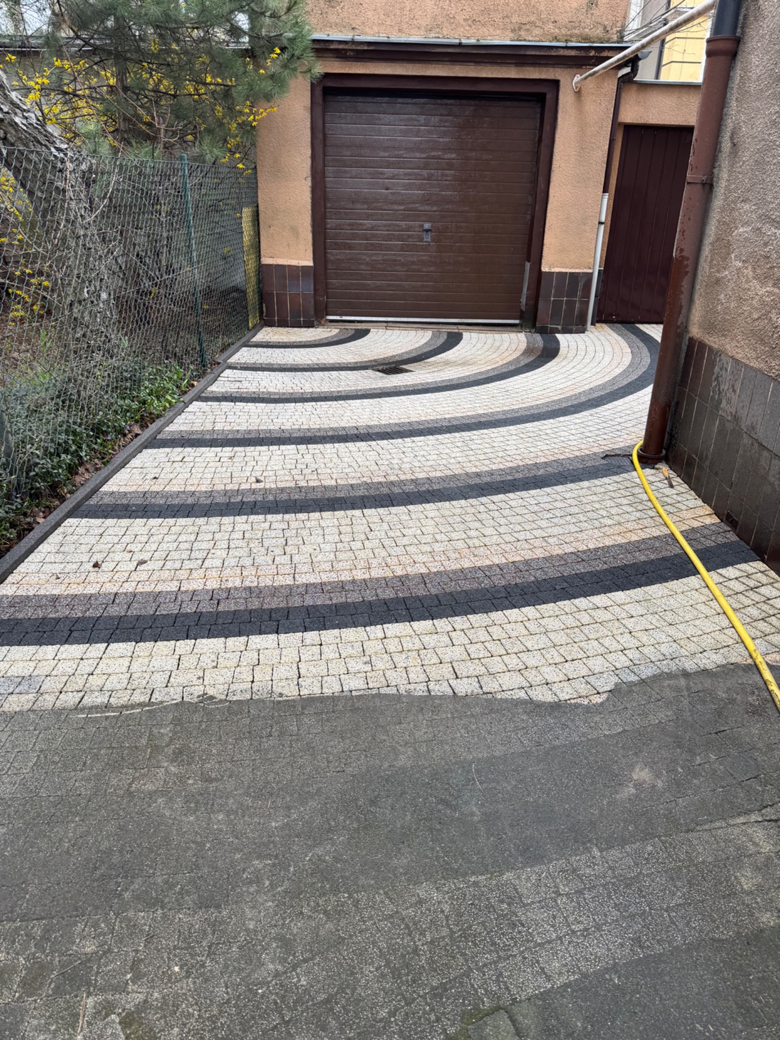 Ułożona w dekoracyjne łuki kostka brukowa przed garażem w Walerianowie, z widocznym fragmentem węża ogrodowego i metalową siatką ogrodzeniową.