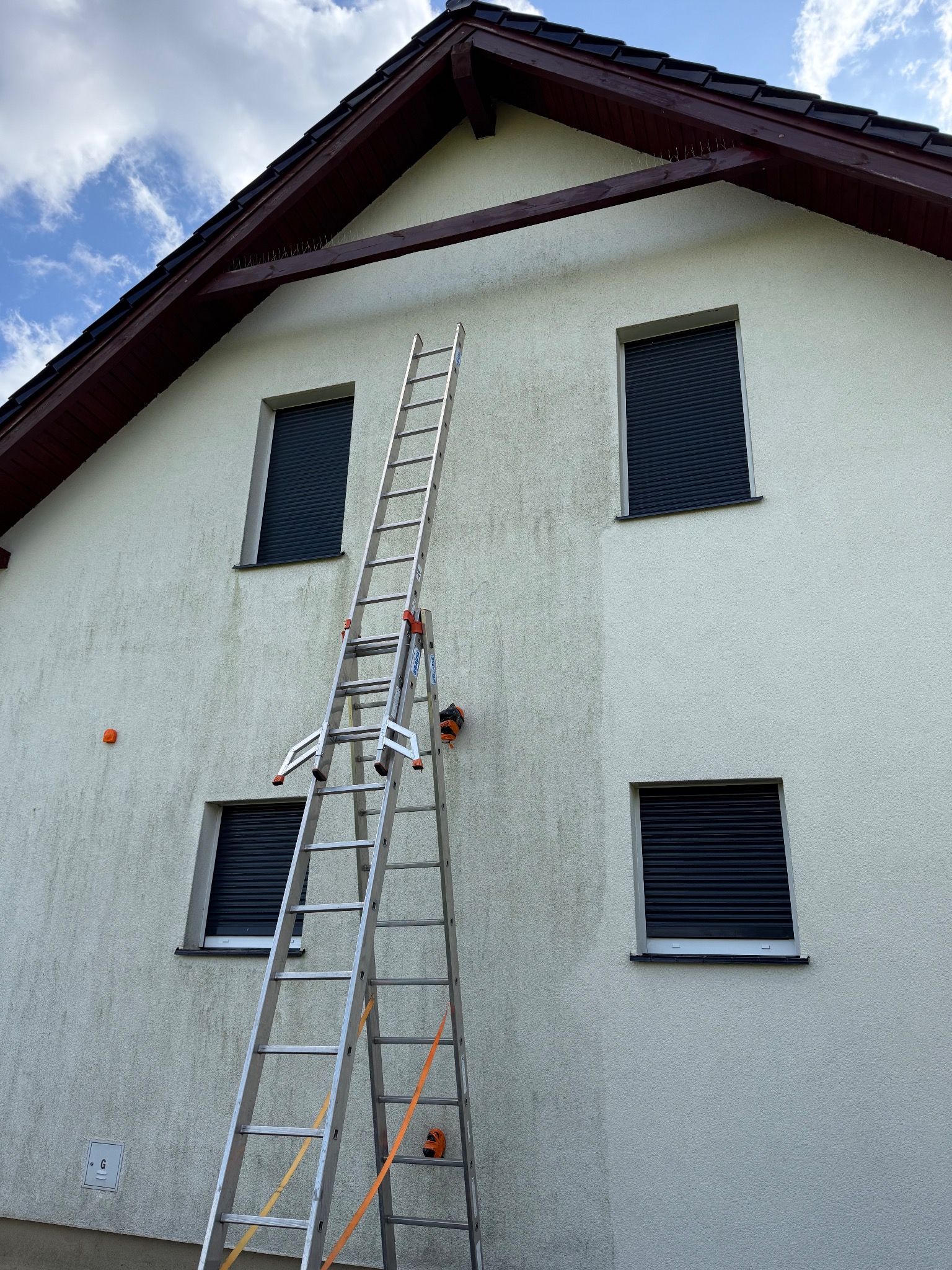 Mycie elewacji domu: widoczny efekt czyszczenia na ścianie, drabina oparta o budynek, zamknięte rolety okienne, błękitne niebo z chmurami w tle.