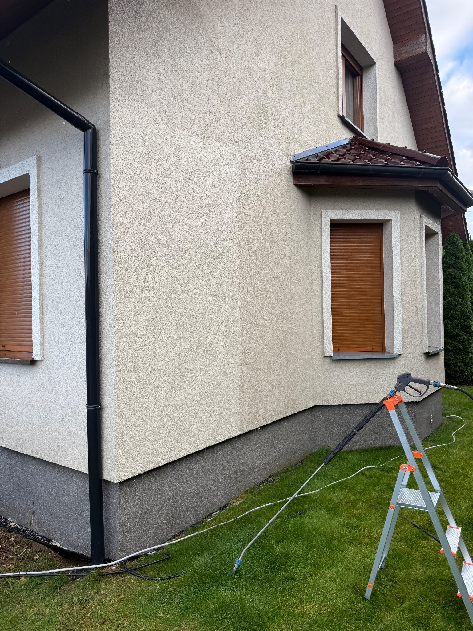 Fragment elewacji domu z widoczną różnicą w czystości po myciu ciśnieniowym. W tle drabina i trawnik, okna z brązowymi roletami.