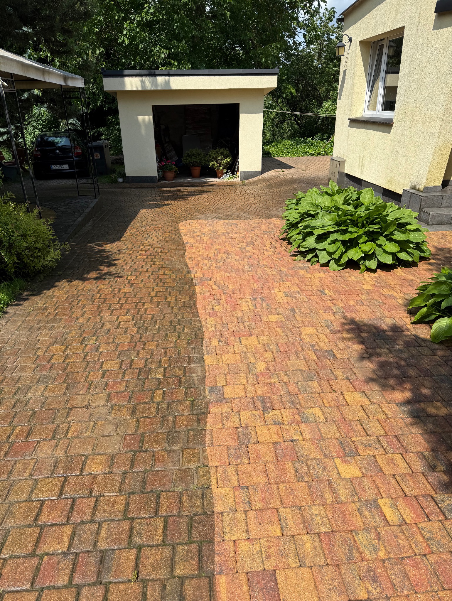 Częściowo umyta, brudna kostka brukowa w odcieniach czerwieni i żółci, z widoczną różnicą czystości. W tle budynek i zieleń.