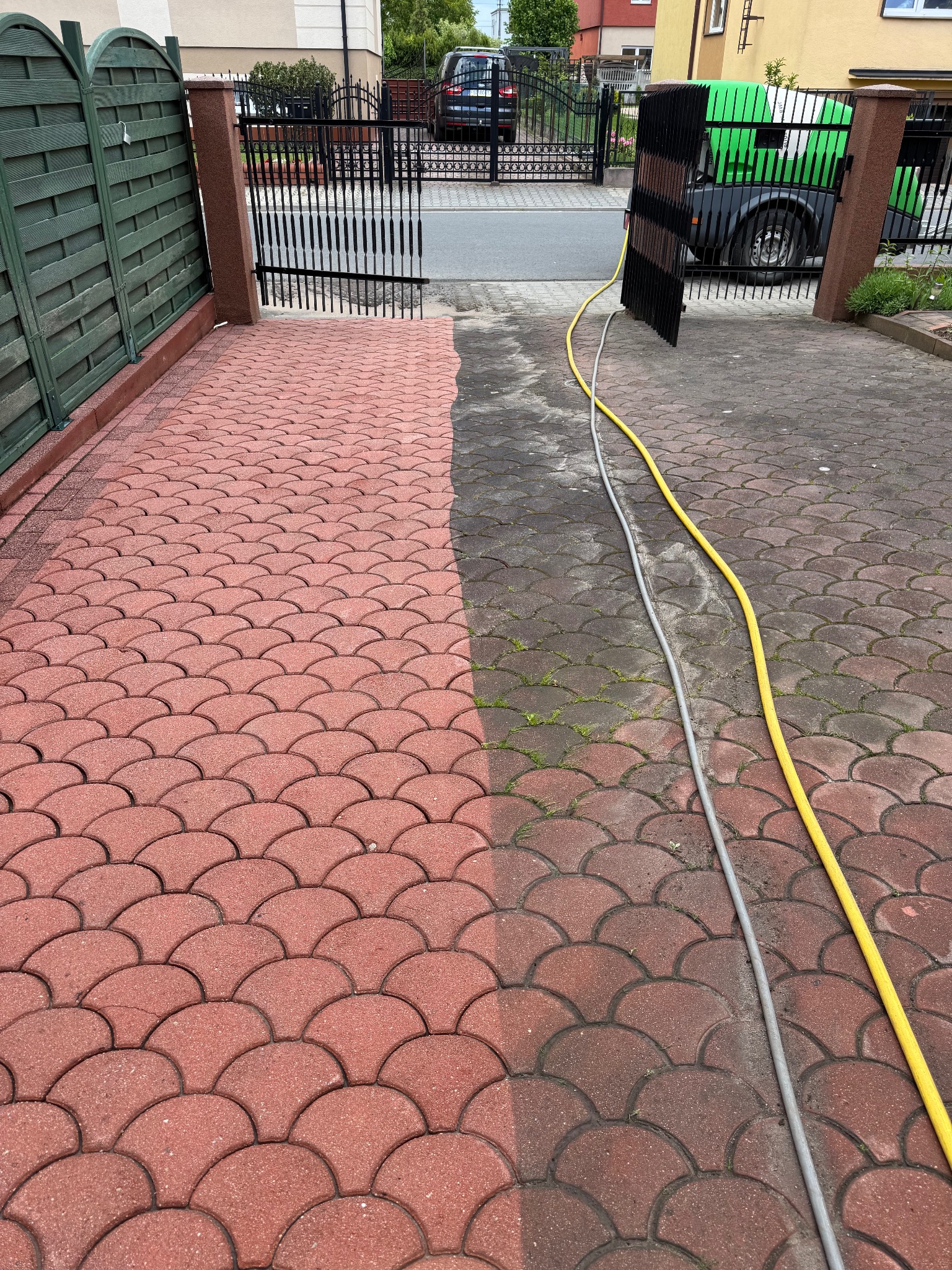 Częściowo wyczyszczona, czerwona kostka brukowa na podjeździe, widoczne węże doprowadzające wodę. Połowa powierzchni jest czysta, druga pokryta zabrudzeniami.