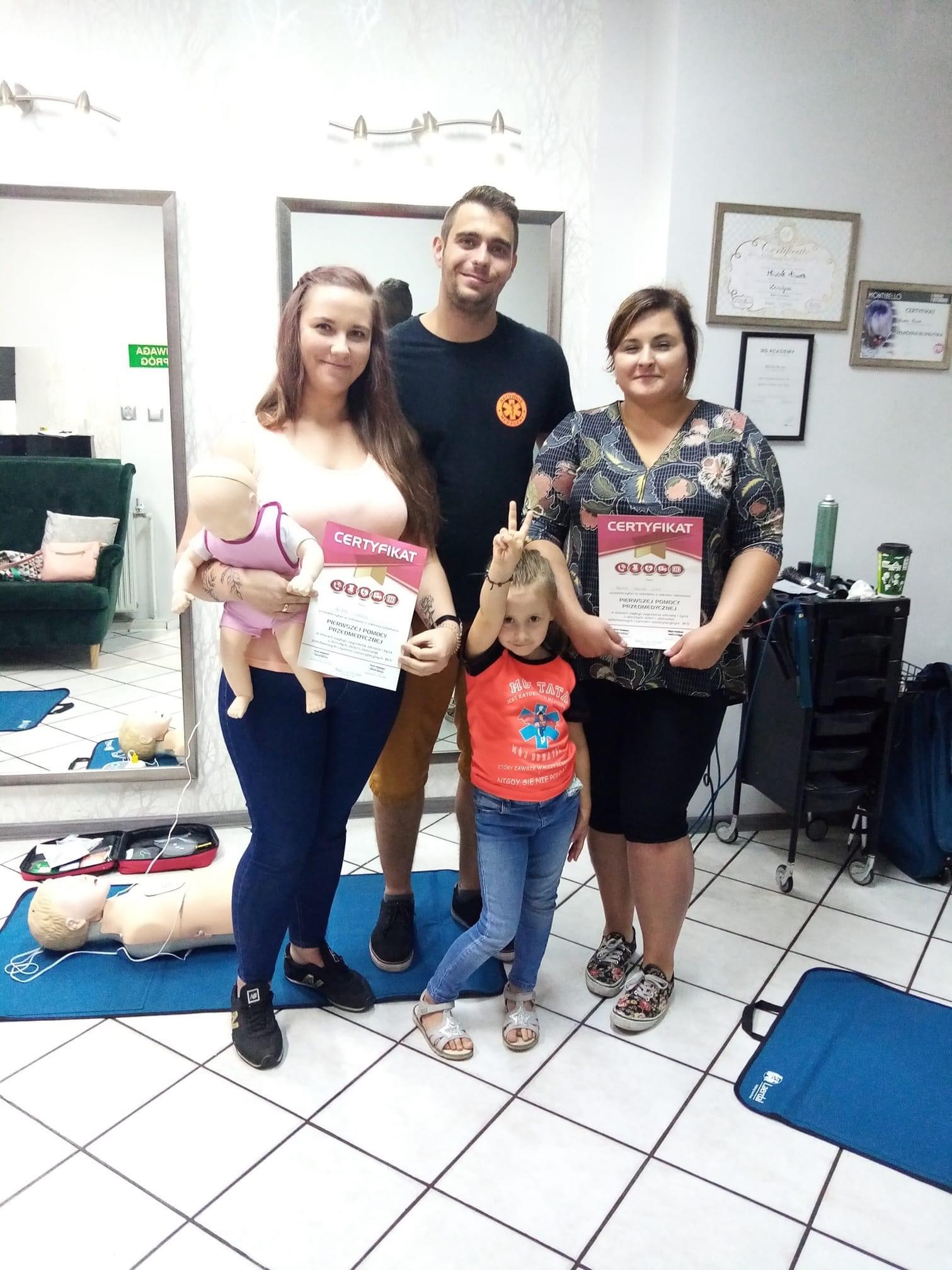 Grupa osób, w tym dziecko, pozuje z certyfikatami ukończenia kursu pierwszej pomocy, obok manekinów do ćwiczeń resuscytacji, w jasnym pomieszczeniu.