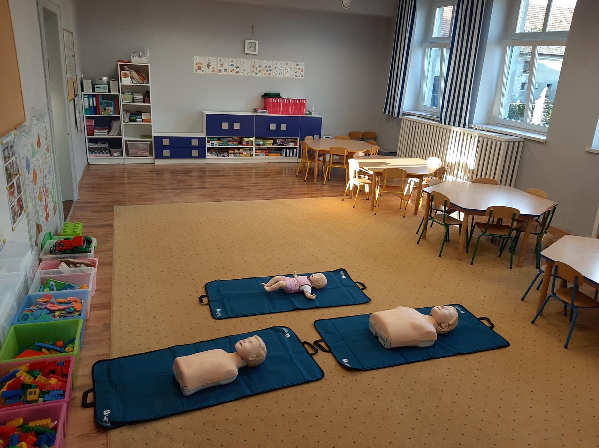 Sala przedszkolna z manekinami do ćwiczeń resuscytacji, w tym jeden manekin niemowlęcia w ubranku, leżące na niebieskich matach.