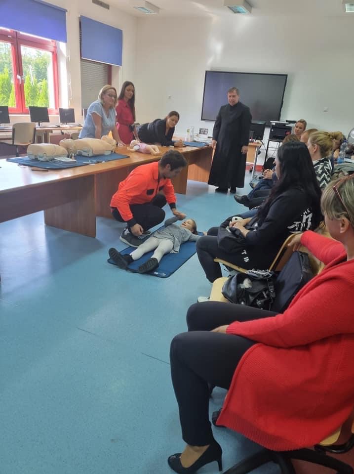 Instruktor w pomarańczowej bluzie demonstruje resuscytację krążeniowo-oddechową na dziecku leżącym na niebieskiej macie, podczas gdy grupa dorosłych obserwuje, a w tle widoczny duchowny w sutannie...