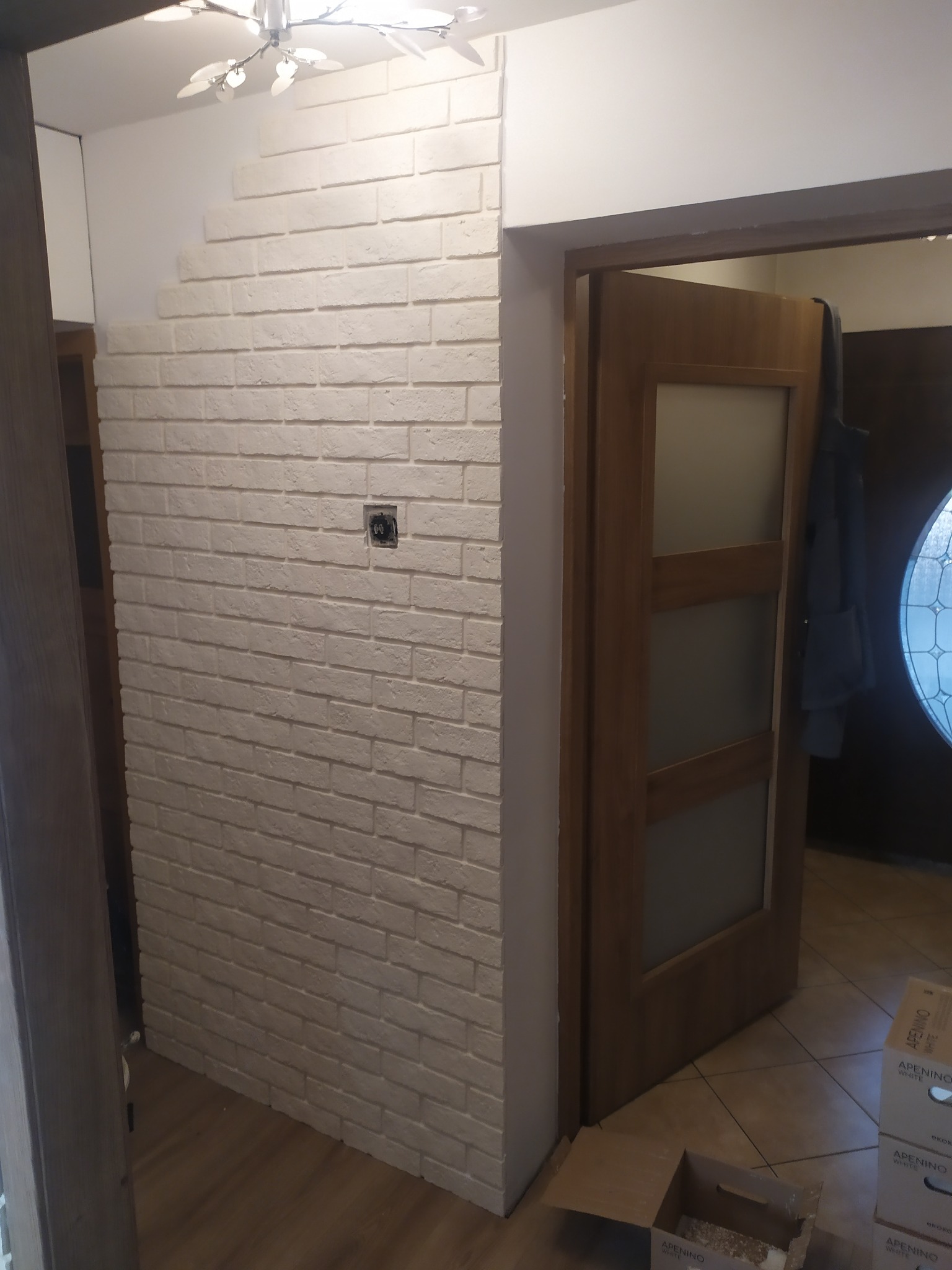 Ściana wykończona białą cegłą dekoracyjną z widocznym gniazdkiem elektrycznym, obok drewniane drzwi wewnętrzne z szybami mlecznymi i kartony z materiałem wykończeniowym na podłodze.
