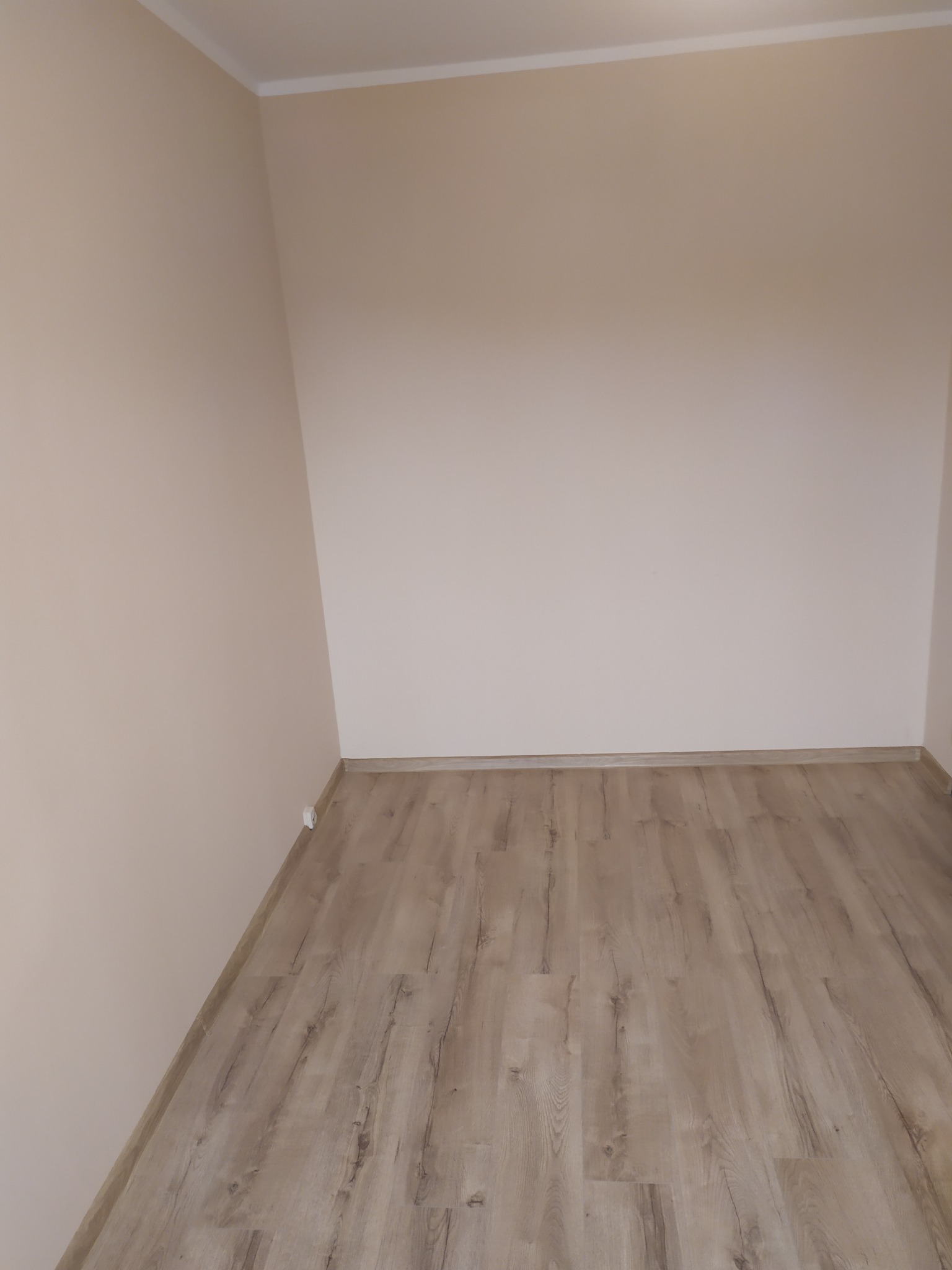 Pusty pokój po remoncie z panelami podłogowymi imitującymi drewno i świeżo pomalowanymi ścianami w odcieniach beżu, widoczne listwy przypodłogowe i sufitowe.