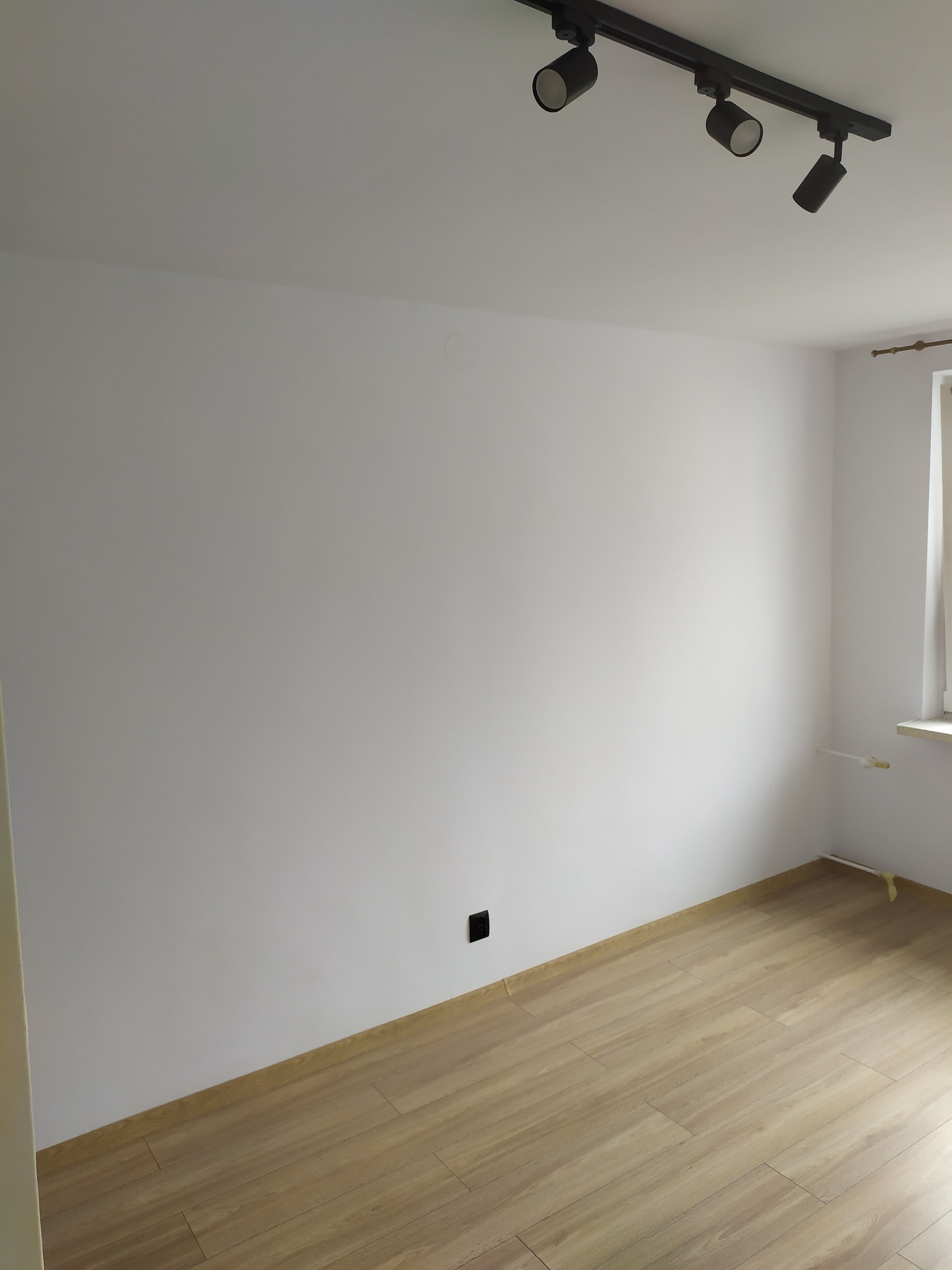 Pusty, odnowiony pokój z jasnymi ścianami, panelami podłogowymi imitującymi drewno i czarną listwą oświetleniową na suficie.