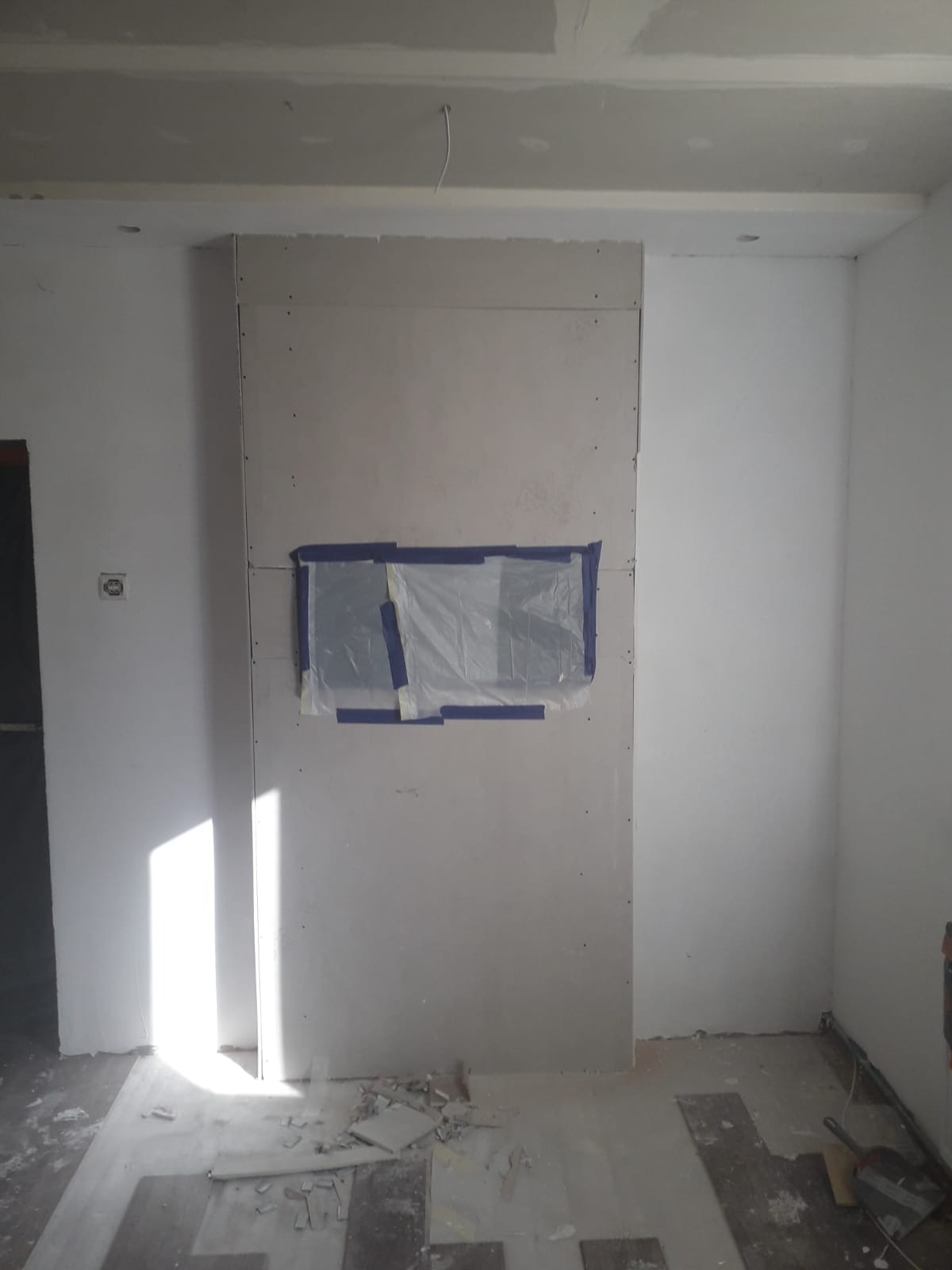Montaż płyty gipsowo-kartonowej na ścianie, z zabezpieczonym otworem folią i taśmą malarską, widoczne ślady po pracy na podłodze.