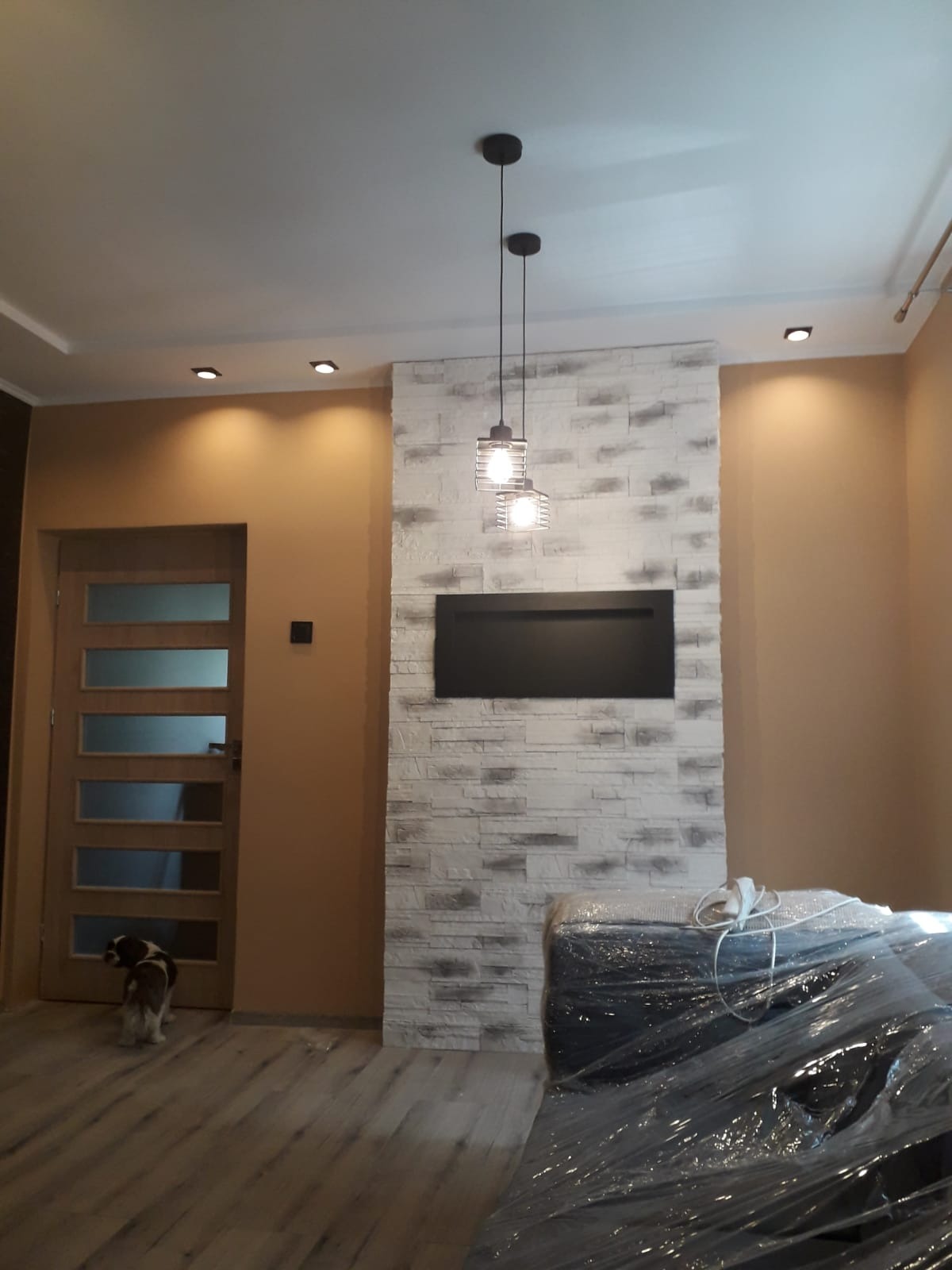 Salon z dekoracyjną ścianą z białej cegły, podwieszonym czarnym panelem i trzema wiszącymi lampami w stylu industrialnym, widoczny częściowo zakryty folią mebel i pies przy drzwiach.