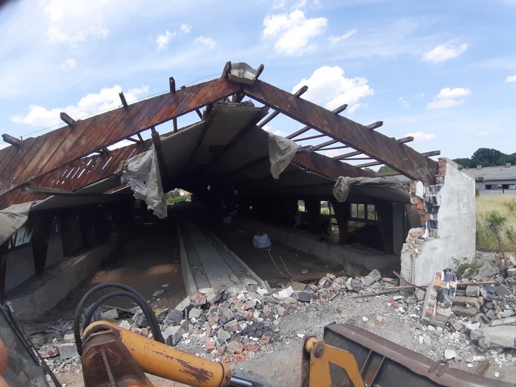 Częściowo zburzony budynek z widoczną konstrukcją dachu i gruzem, fragment maszyny budowlanej w dolnej części kadru.